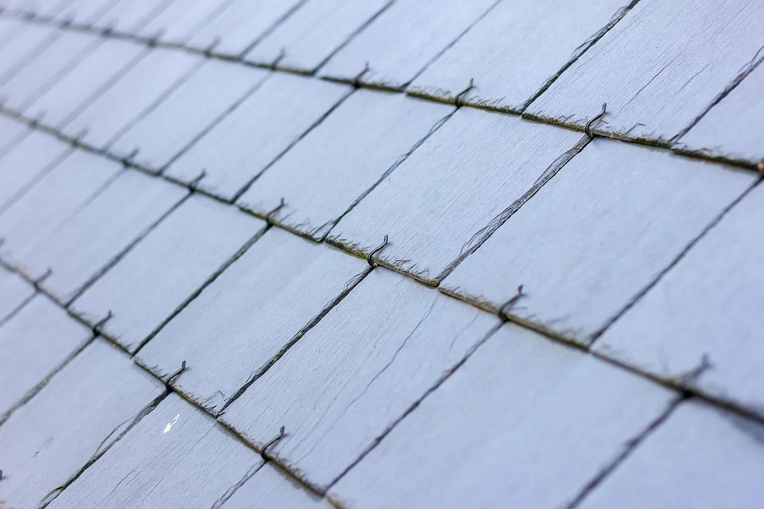 Close-up of blue-gray wooden shingles on a roof, with small metal hooks holding some shingles in place.