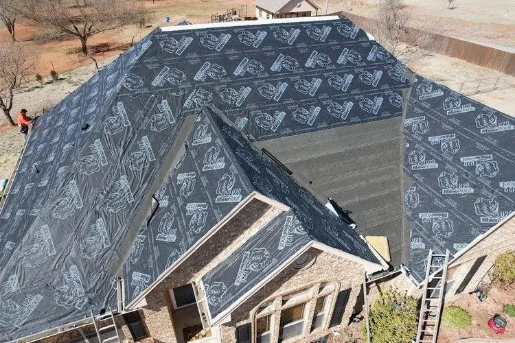 A house under construction with a roof partially covered in black weatherproofing material, surrounded by construction tools, ladders, and workers.