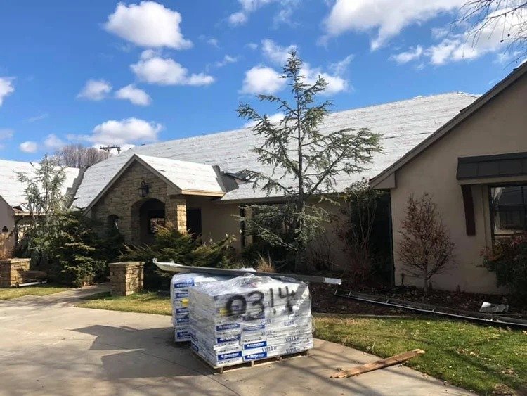 A house with snow-covered roof, trees, and a pallet of construction materials on the driveway.