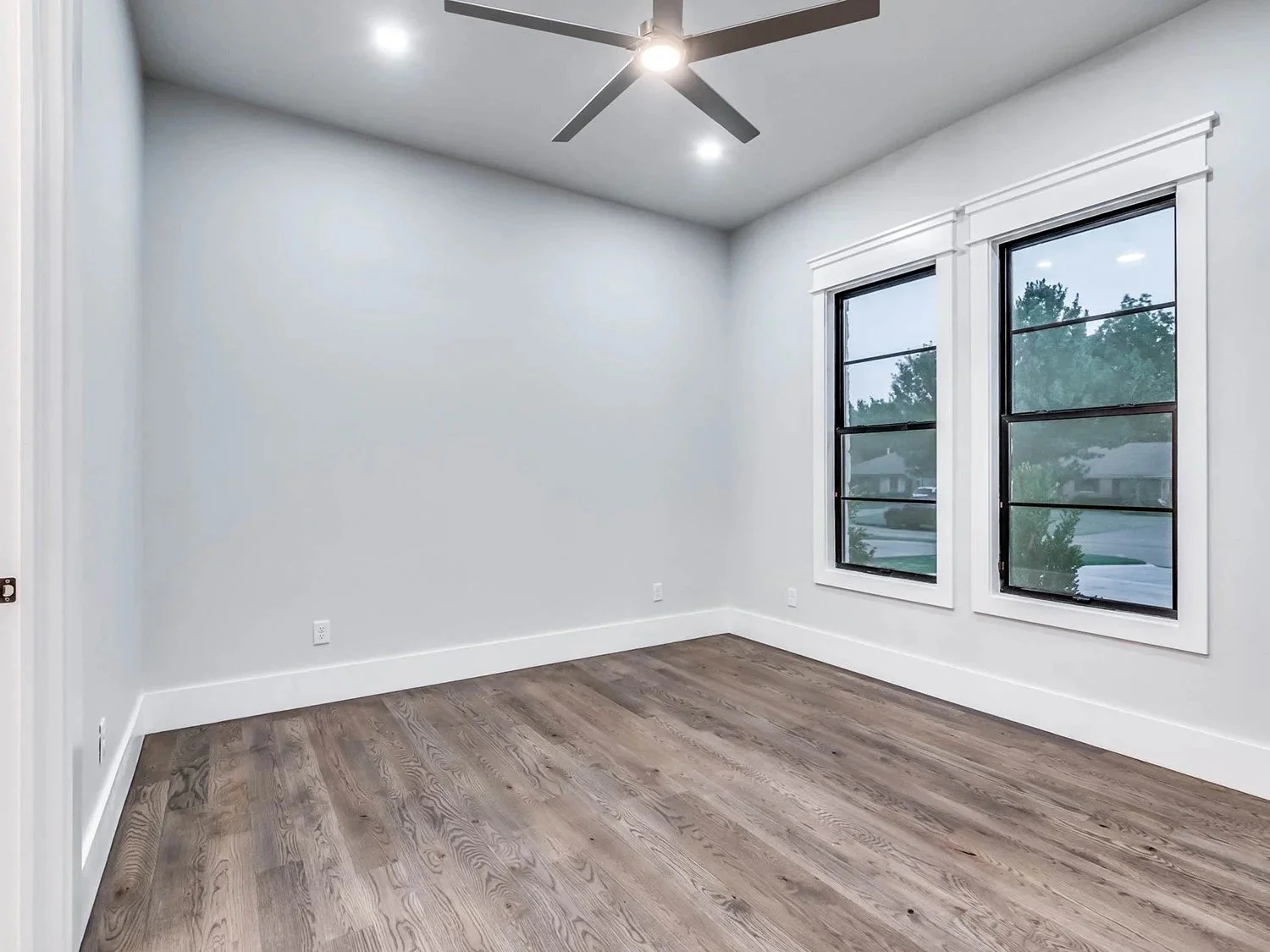 Empty room with white walls, hardwood floor, large windows, and a ceiling fan with lights.