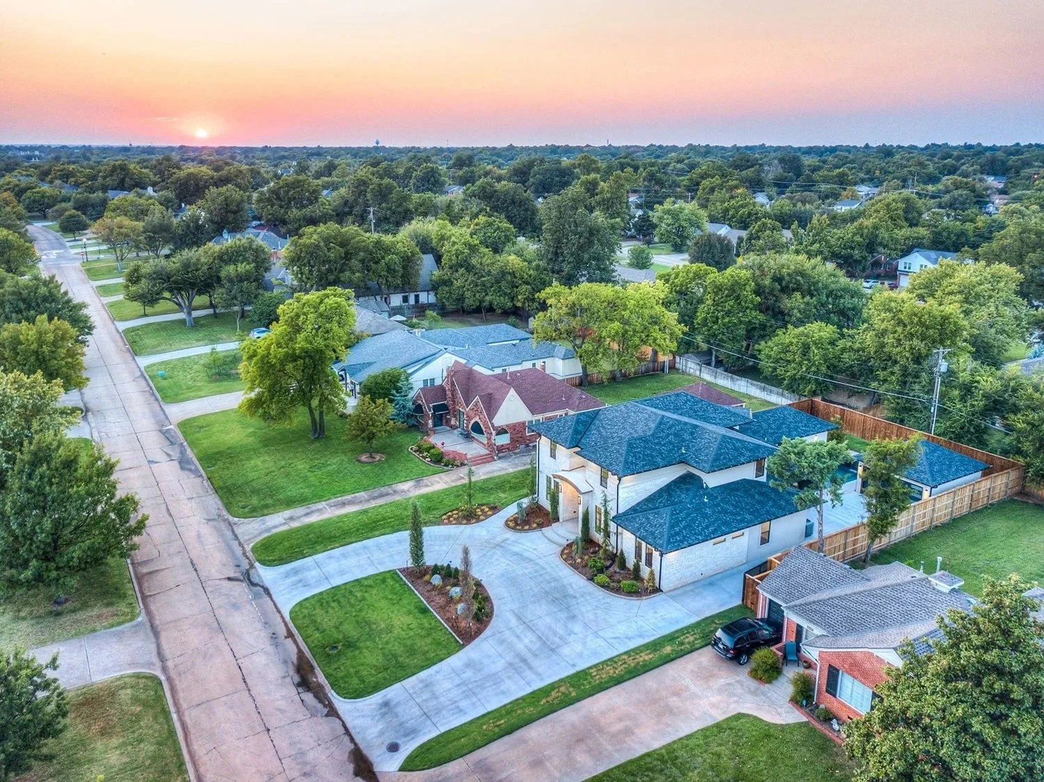 Aerial view of a suburban neighborhood at sunset showing large houses, green lawns, trees, and streets.
