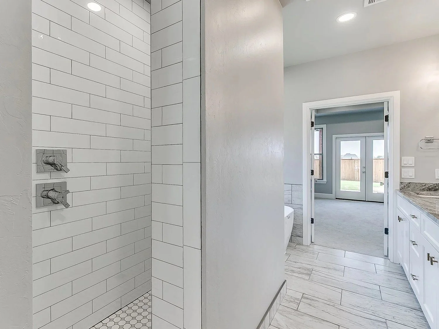 Bathroom with walk-in shower featuring white subway tiles and hexagonal floor tiles, view toward an adjoining area with French doors leading outside, a bathtub, and bathroom cabinetry with granite countertop.