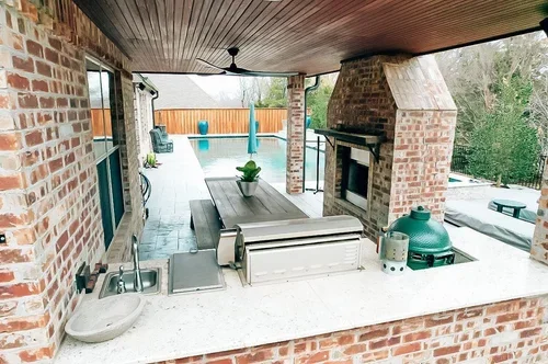 Outdoor kitchen with a brick counter, sink, grill, and fireplace, overlooking a pool with blue umbrellas and lounge chairs.