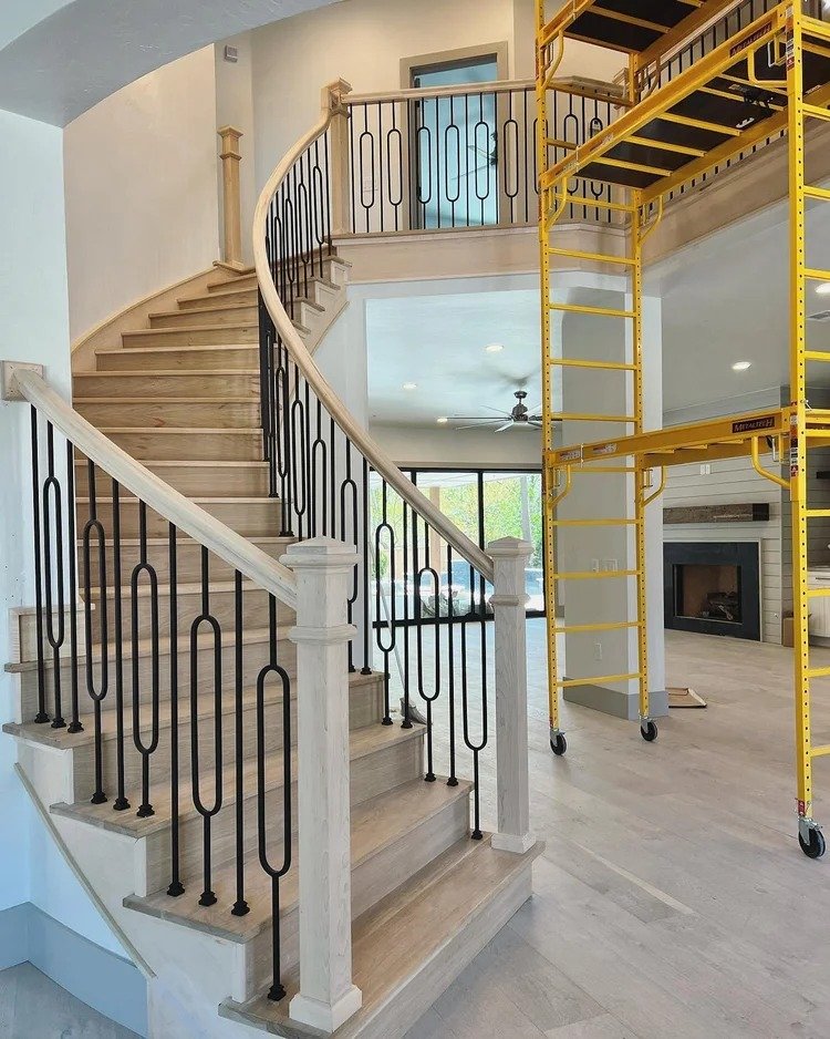 Interior of a house under renovation, showing a curved staircase with light wood steps and black metal railing, alongside a yellow scaffolding structure, in a bright living room with a nearby fireplace and large windows.