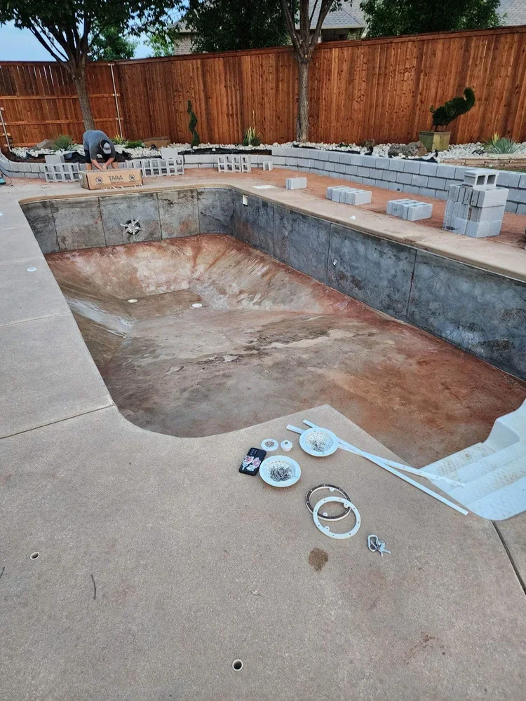 Empty swimming pool under construction with tools and materials around, a worker installing bricks, surrounded by trees and a wooden fence.