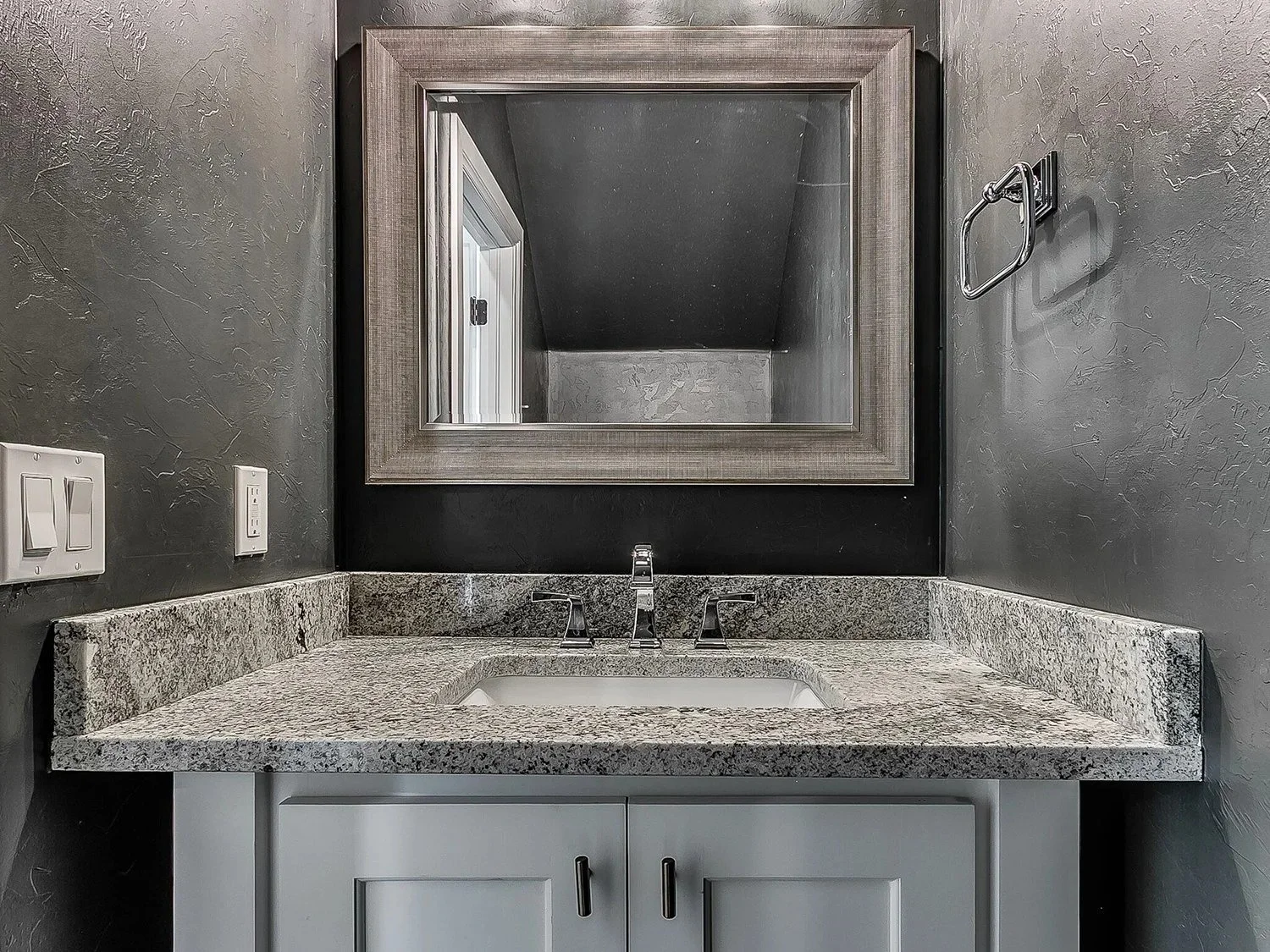 Bathroom vanity with granite countertop, white cabinets, large framed mirror, gray textured walls, faucet, electrical outlets, and a vacant towel ring.