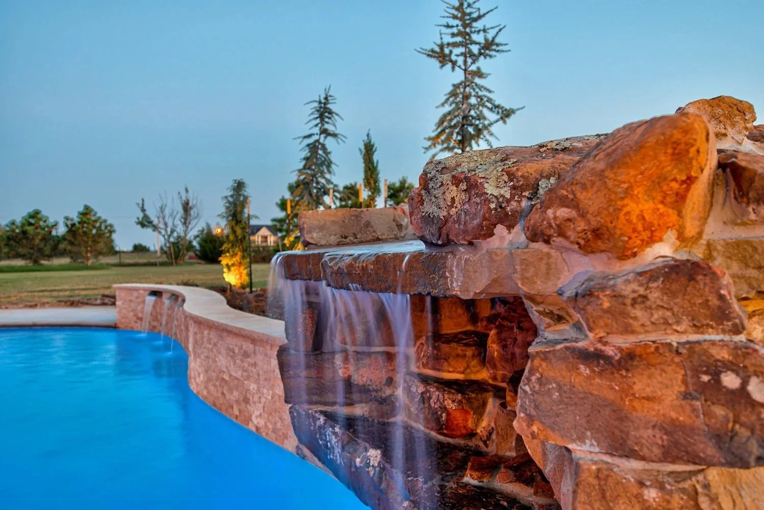Backyard swimming pool with a waterfall feature made of natural rocks, surrounded by greenery and trees during dusk or evening.