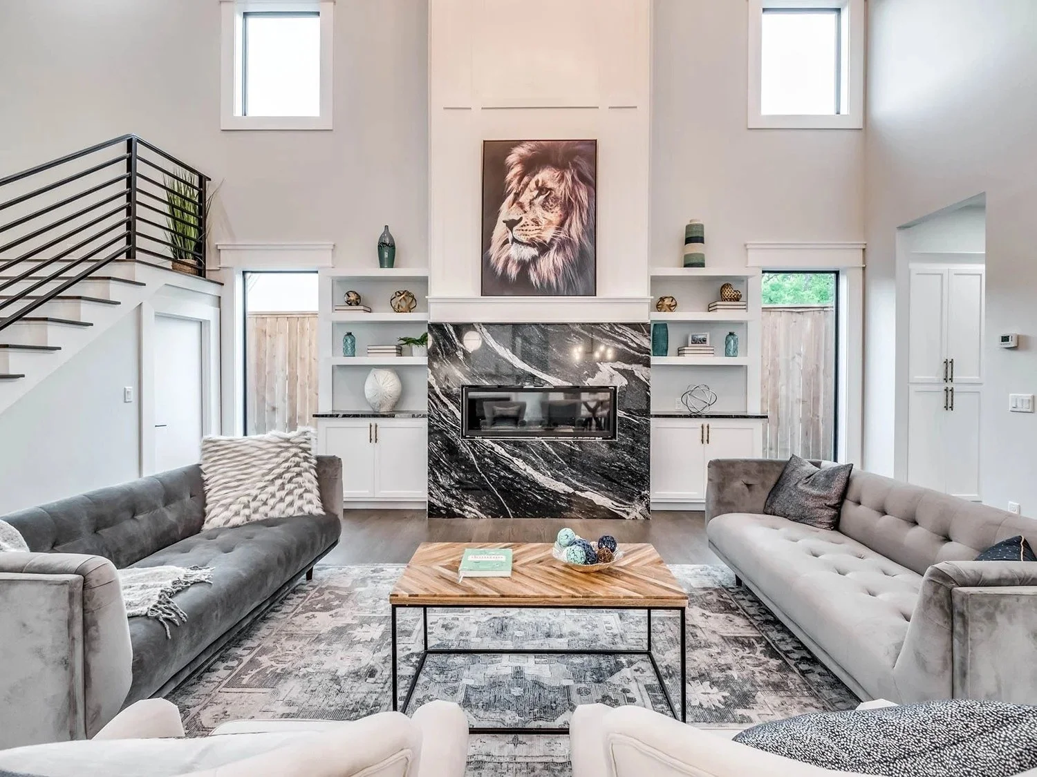 Living room with two gray sofas, wooden coffee table, fireplace with black and white marble, built-in shelves with decorative items, large white walls, and multiple windows, featuring a lion portrait above the fireplace.