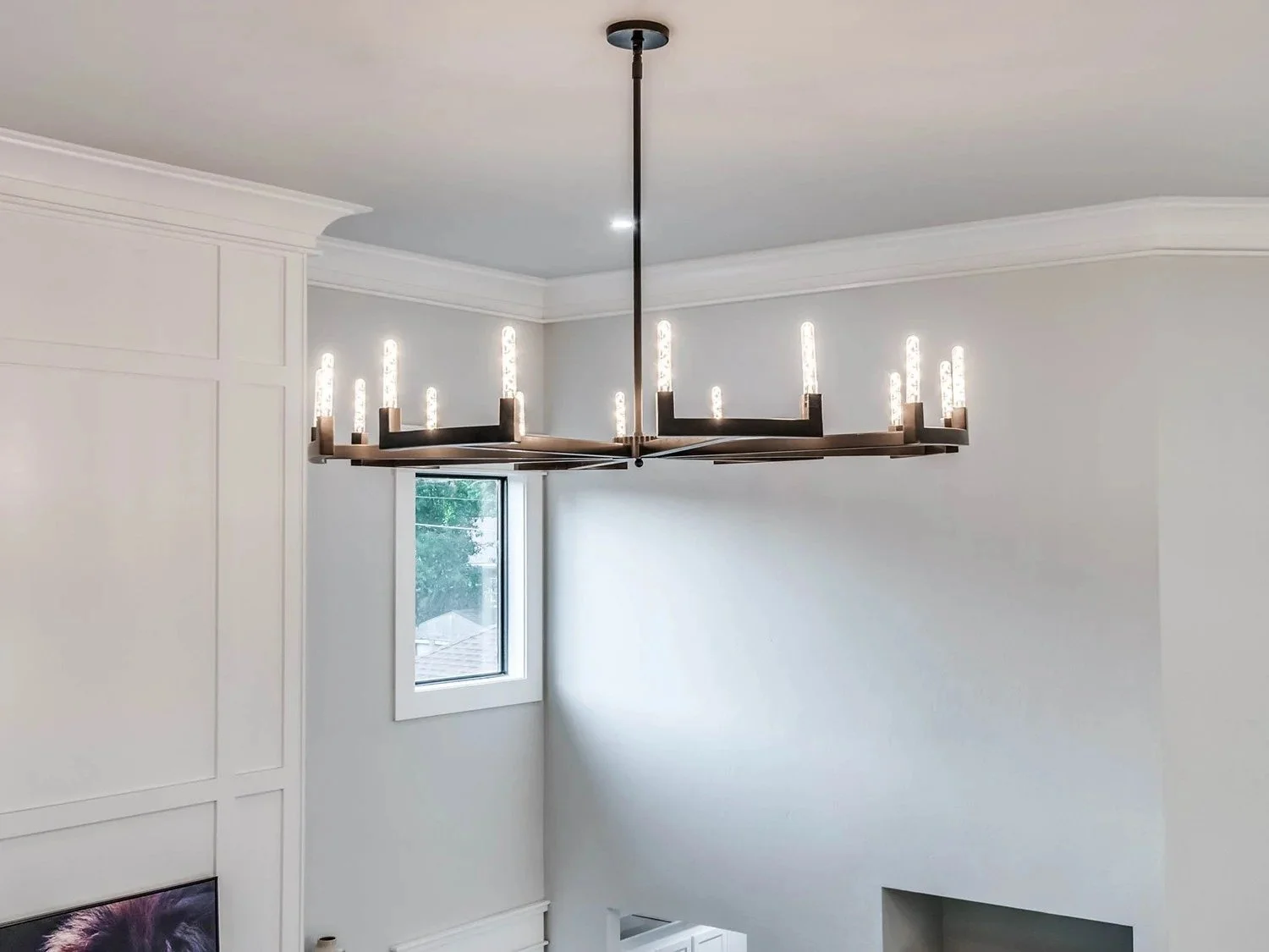 Modern black chandelier with exposed bulbs hanging from the ceiling in a white room with crown molding and a small window.