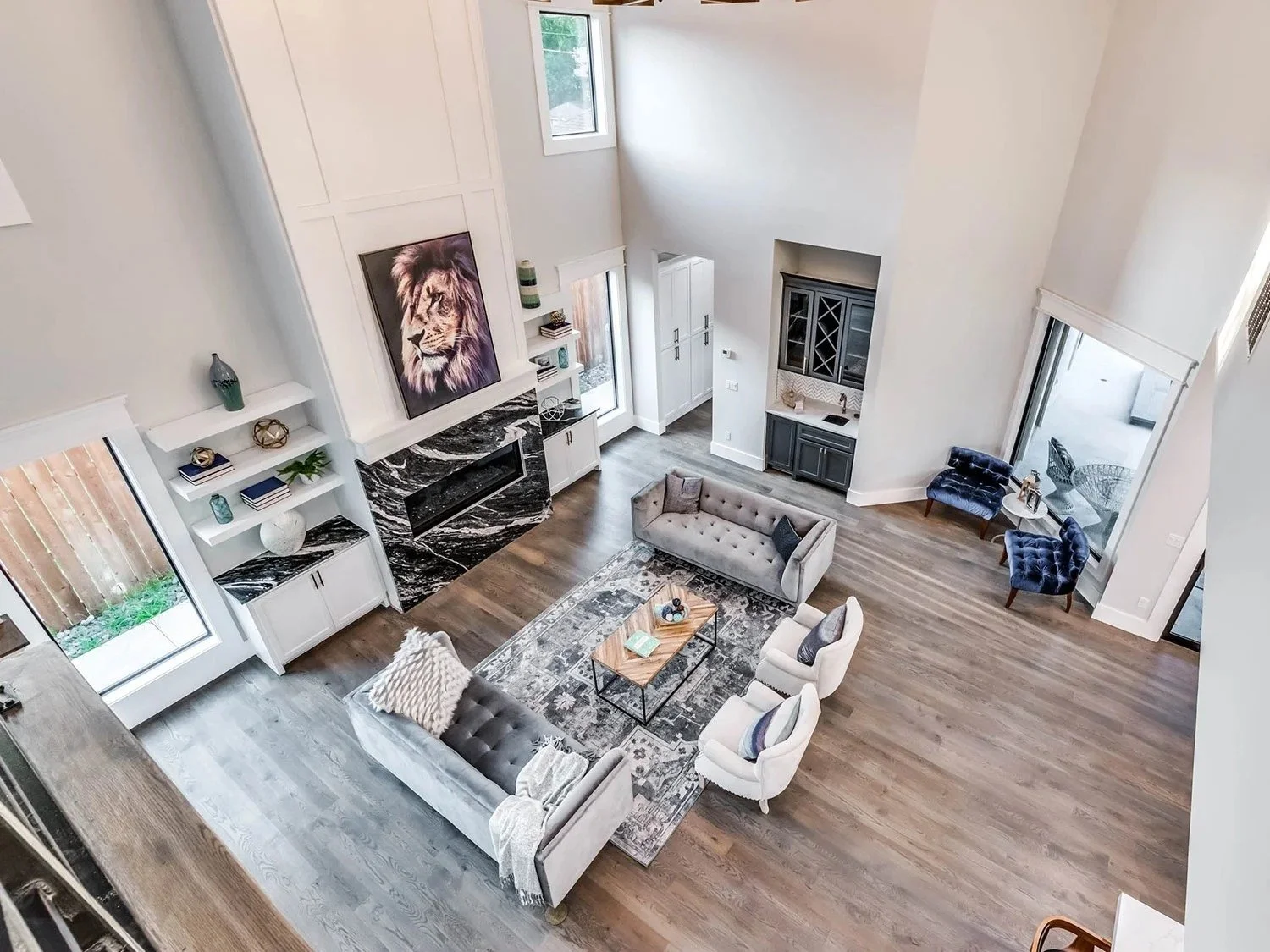 A spacious, modern living room with high ceilings, large windows, white walls, and hardwood floors. Includes a grey sofa, white armchairs, a black and white patterned rug, a fireplace with a black marble surround, and artwork featuring a lion. There 
