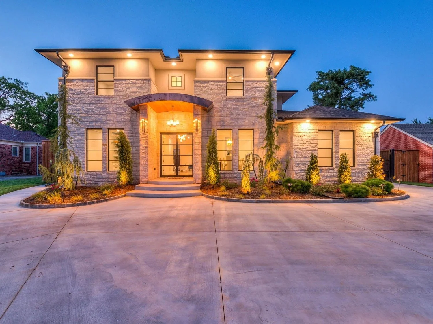 A modern two-story house with stone exterior, large windows, and a well-lit entrance surrounded by landscaped garden with small trees and shrubs, during twilight.