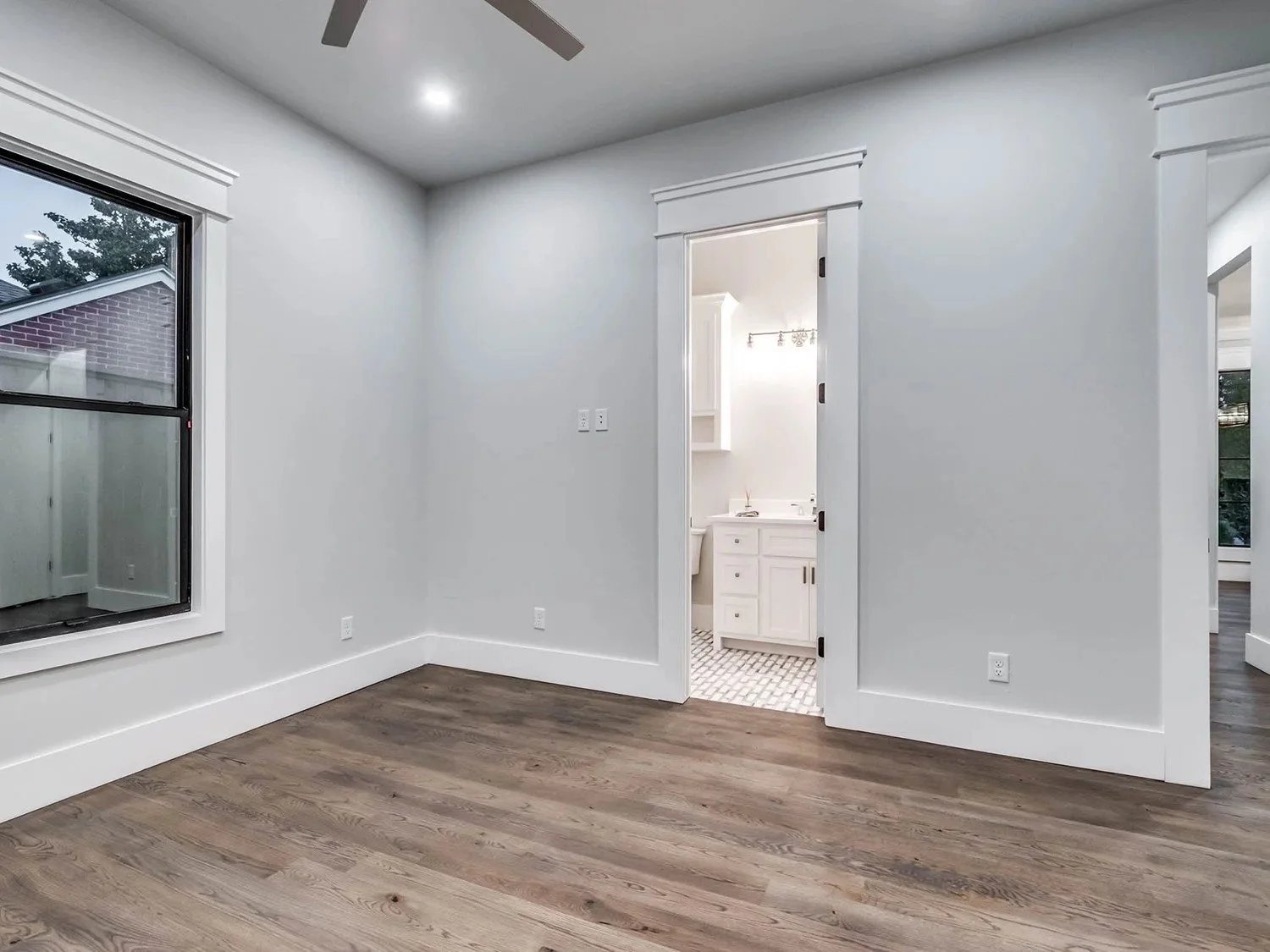 Empty room with hardwood flooring, window, and doorway leading to a bathroom with a white vanity and patterned tile floor.