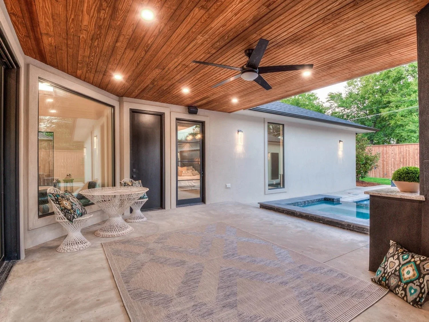 Covered backyard patio with wood-paneled ceiling, ceiling fan, recessed lighting, small pool, seating area with patterned cushions, glass door and windows, and a border fence with green trees in the background.