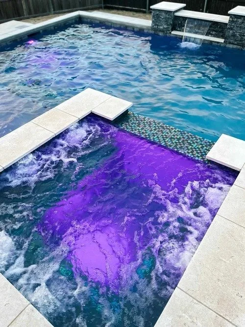 A backyard swimming pool with a smaller hot tub attached, illuminated with purple and blue underwater lighting.