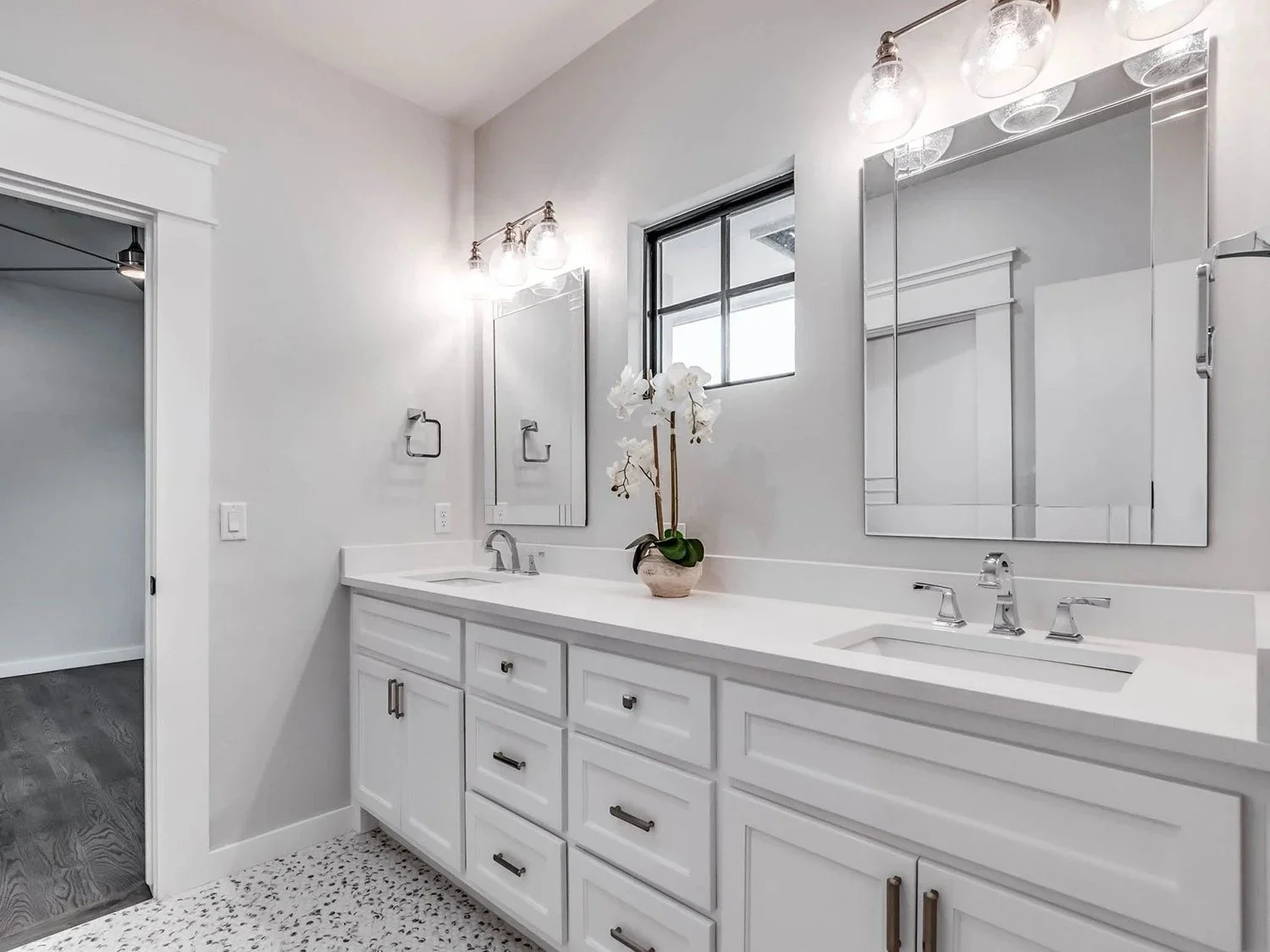 Modern bathroom with white cabinetry, dual sinks, large mirrors, wall-mounted light fixtures, a window, and a potted orchid