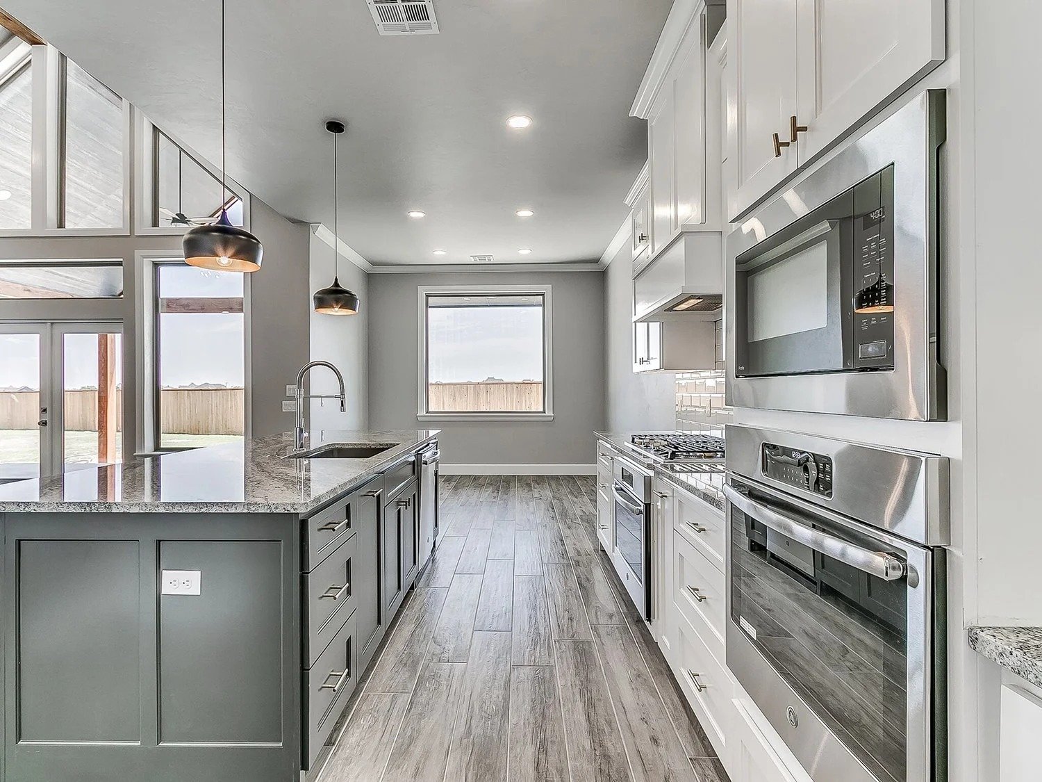 Modern kitchen with gray cabinets, stainless steel appliances, granite countertops, a large window, wooden flooring, and pendant lighting.