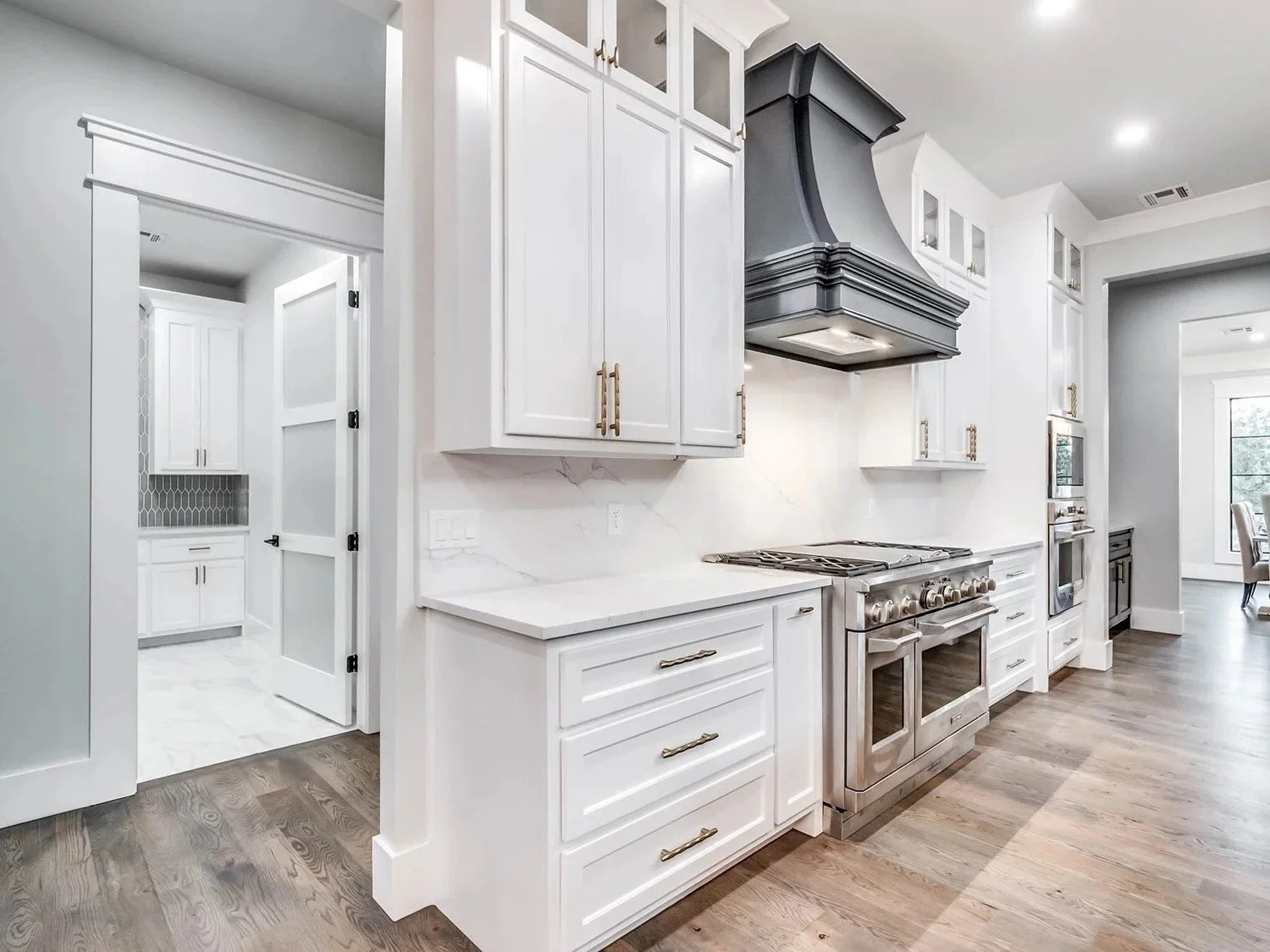 Modern kitchen with white cabinets, stainless steel oven, black range hood, and wooden flooring.