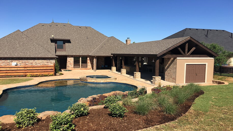 Backyard with a kidney-shaped swimming pool, patio, covered pavilion, brick house, and landscaped garden.
