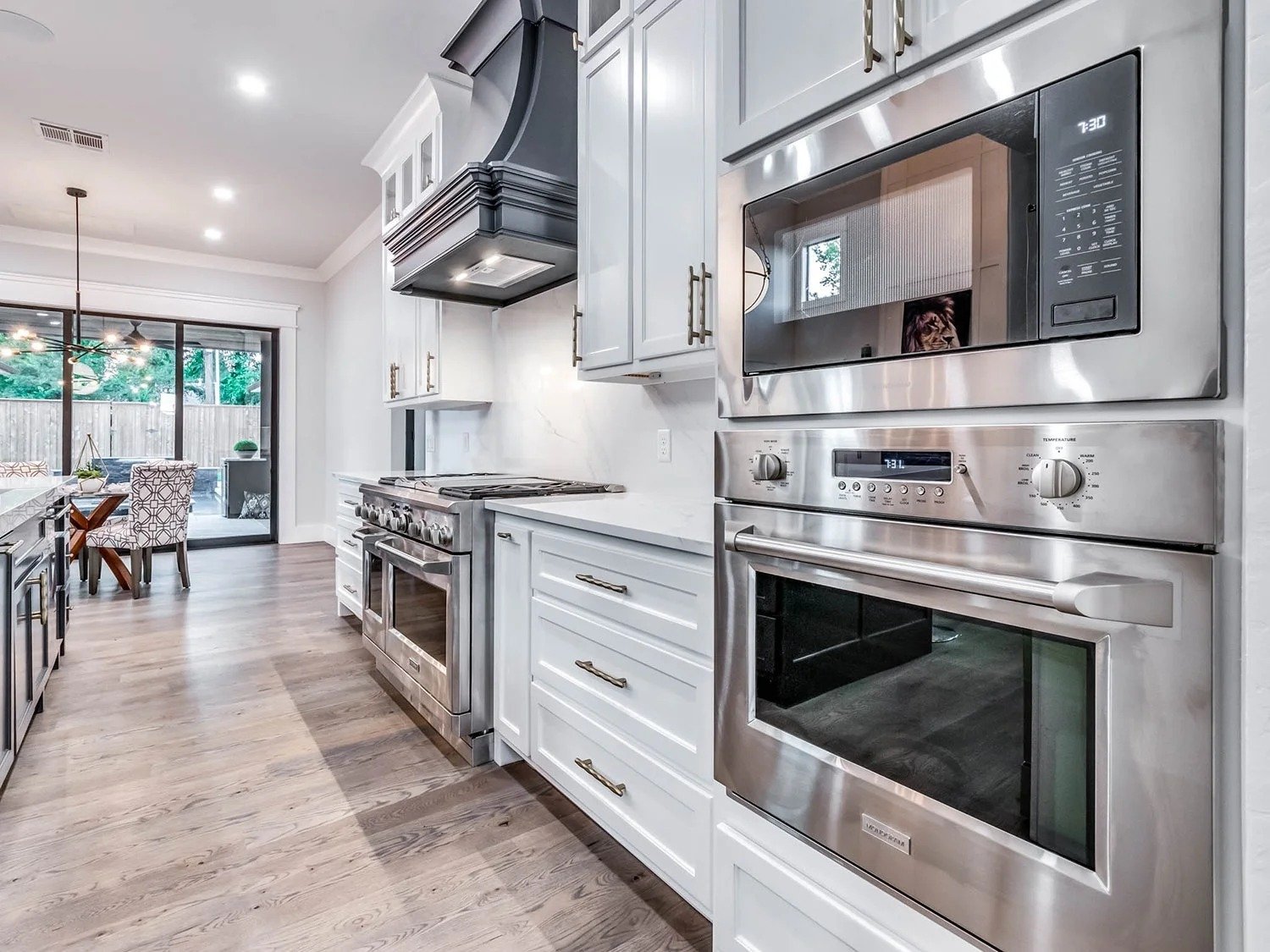 Modern kitchen with white cabinets, stainless steel appliances including microwave, oven, and stove, and a view of a dining area with sliding glass doors leading to an outdoor space.