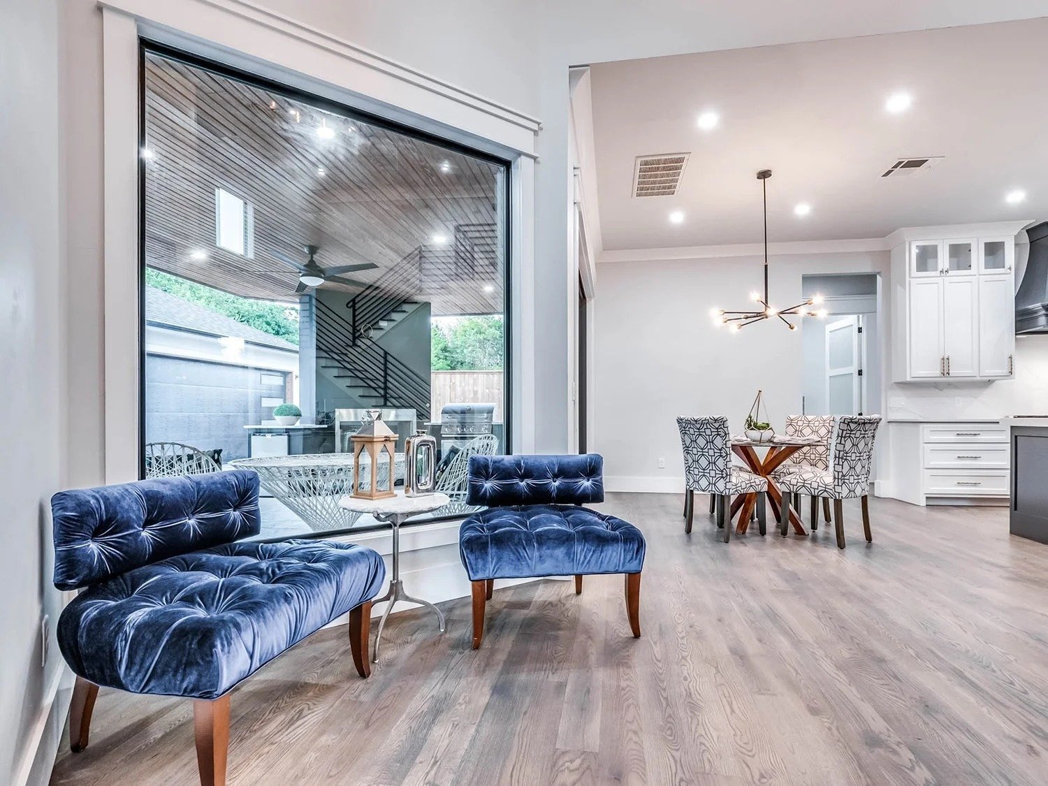 Interior of a modern living area with blue velvet settees, a glass side table, a dining table with patterned chairs, hardwood flooring, white cabinets, and a large window showing an outdoor patio with stairs, ceiling fan, and greenery.