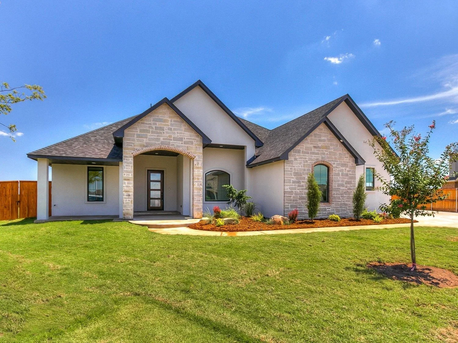 Newly built modern house with stone and stucco exterior, front yard with green lawn, small trees, and landscaping, under a clear blue sky.
