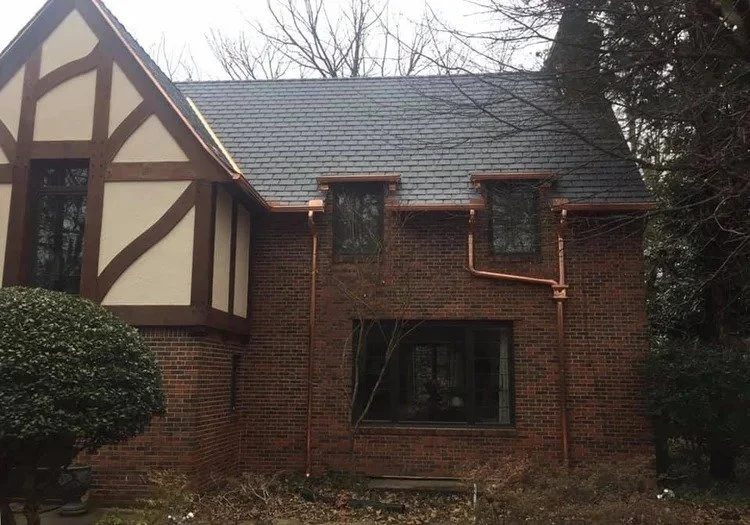 A brick house with a slate roof, featuring a front window and two small upper windows, surrounded by trees and bushes.