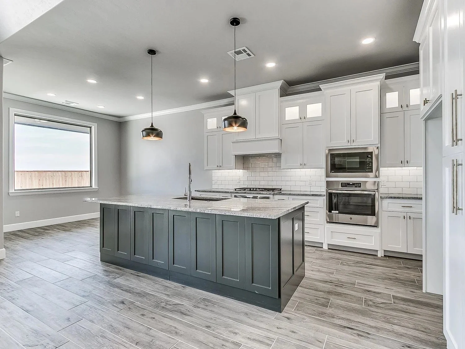 A modern kitchen with a large island with a grey base and a white marble countertop, white cabinets, stainless steel appliances, wood-look tile flooring, a window, and pendant lights hanging from the ceiling.