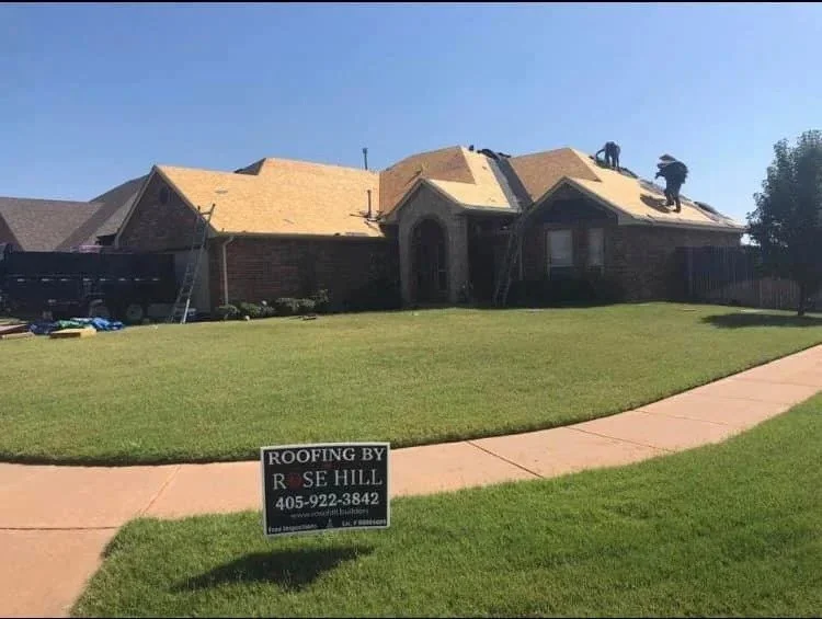 A house with roof work in progress, two workers on the roof, ladders leaning against the house, and a sign in the lawn advertising roofing services.