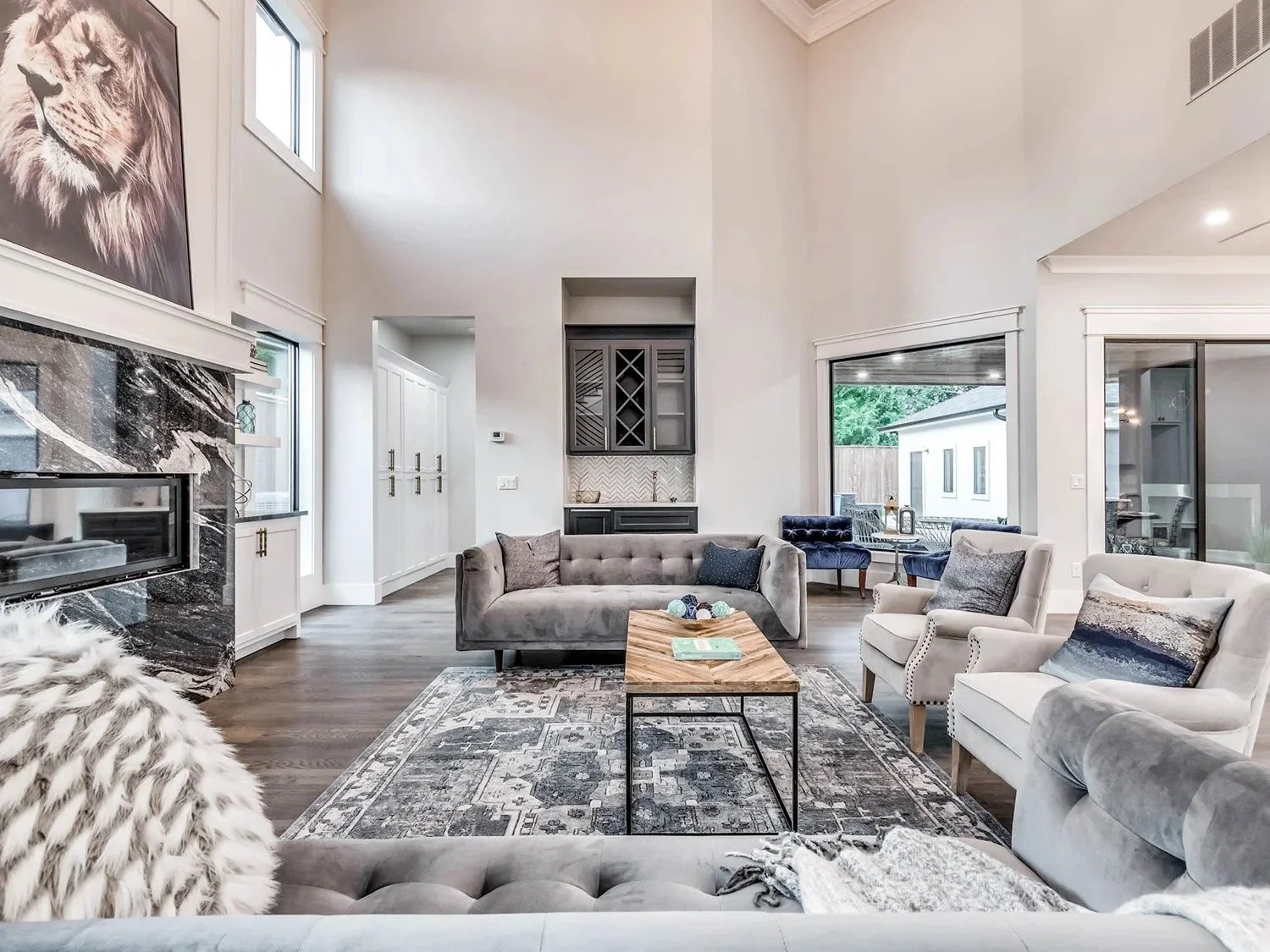 Spacious living room with high ceilings, featuring white walls, large windows, and modern furniture including gray and cream armchairs, a gray sofa, and a wooden coffee table. Decor includes a patterned area rug, pillows, and a wall-mounted painting 