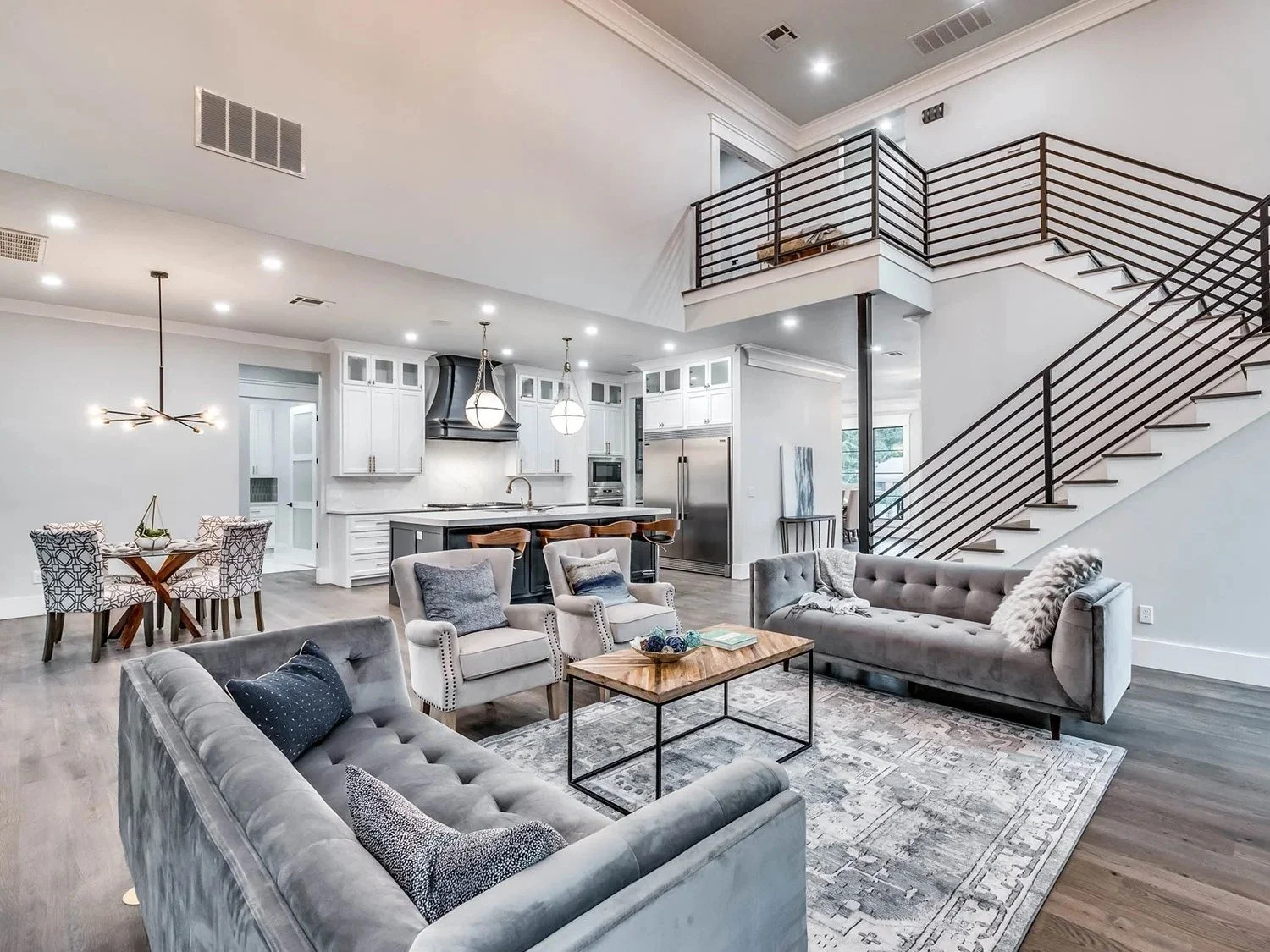 Open plan living room and kitchen with modern decor, gray sofas, a wooden coffee table, a dining table with four chairs, a staircase with black railing, and stainless steel appliances in the kitchen.