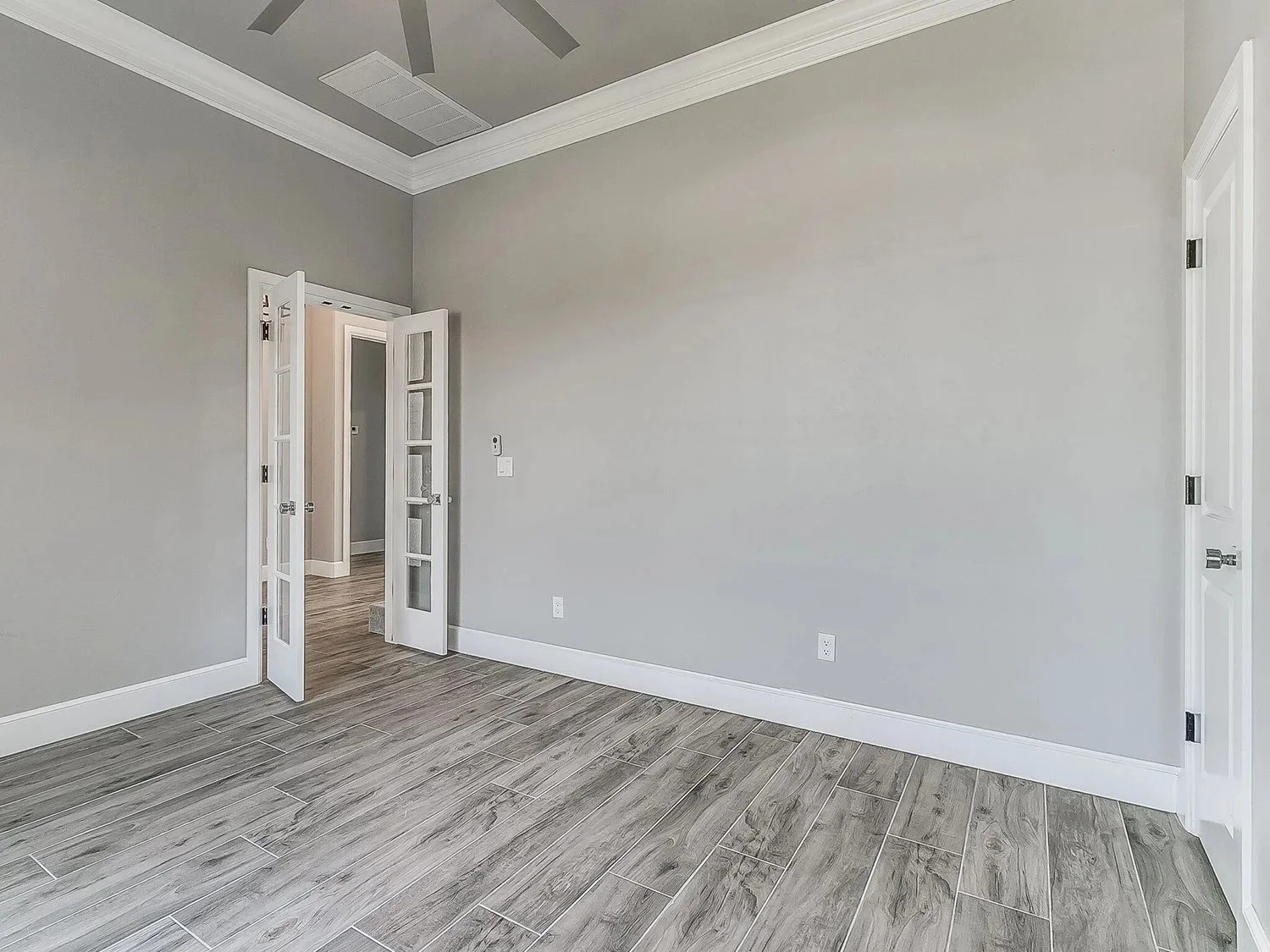 Empty room with gray walls, white trim, and gray wooden flooring. An open door with glass panes leads to another room with similar flooring. Ceiling with vents and crown molding.