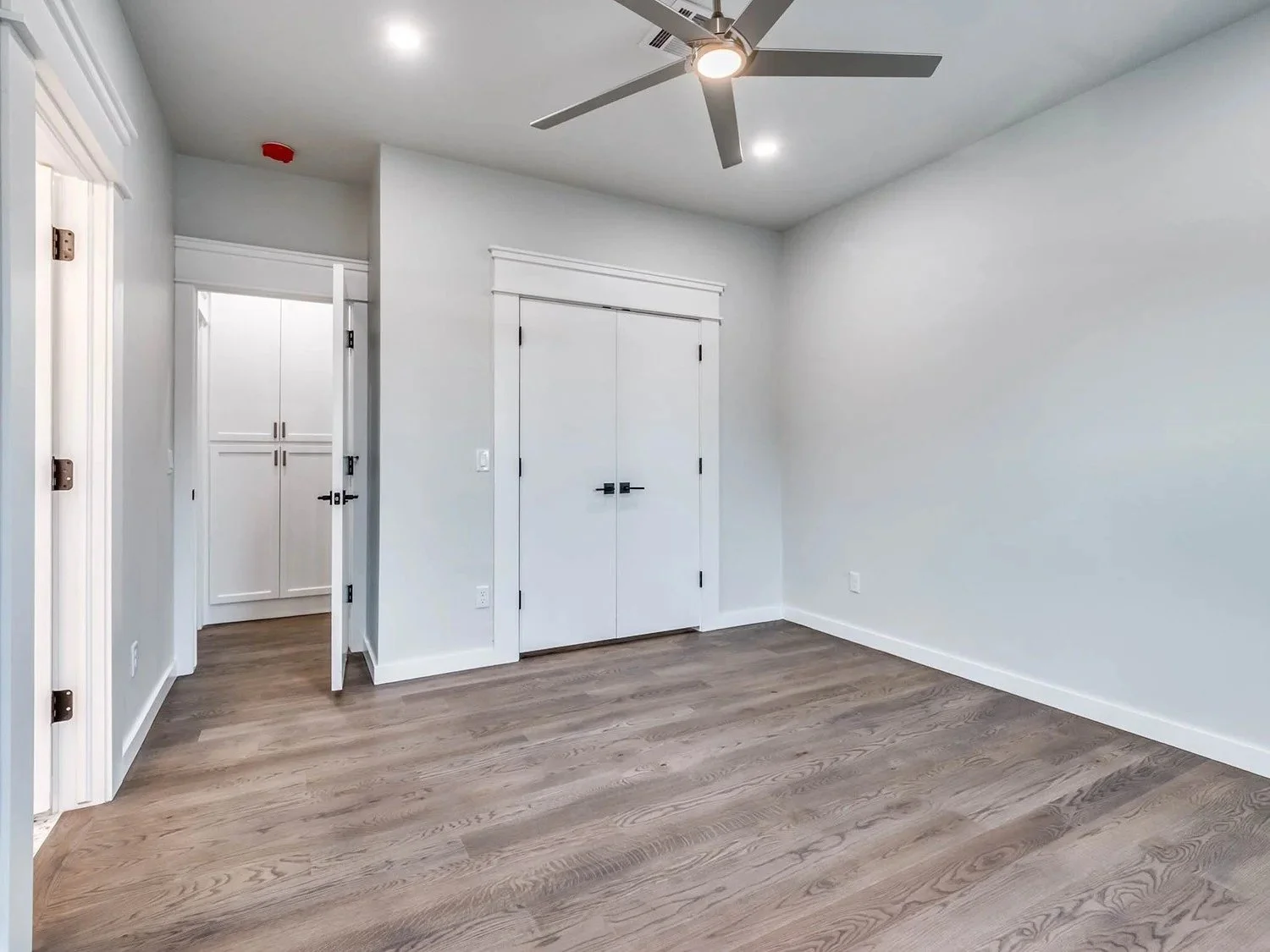 Empty room with hardwood floors, white walls, a ceiling fan, and built-in white cabinets.