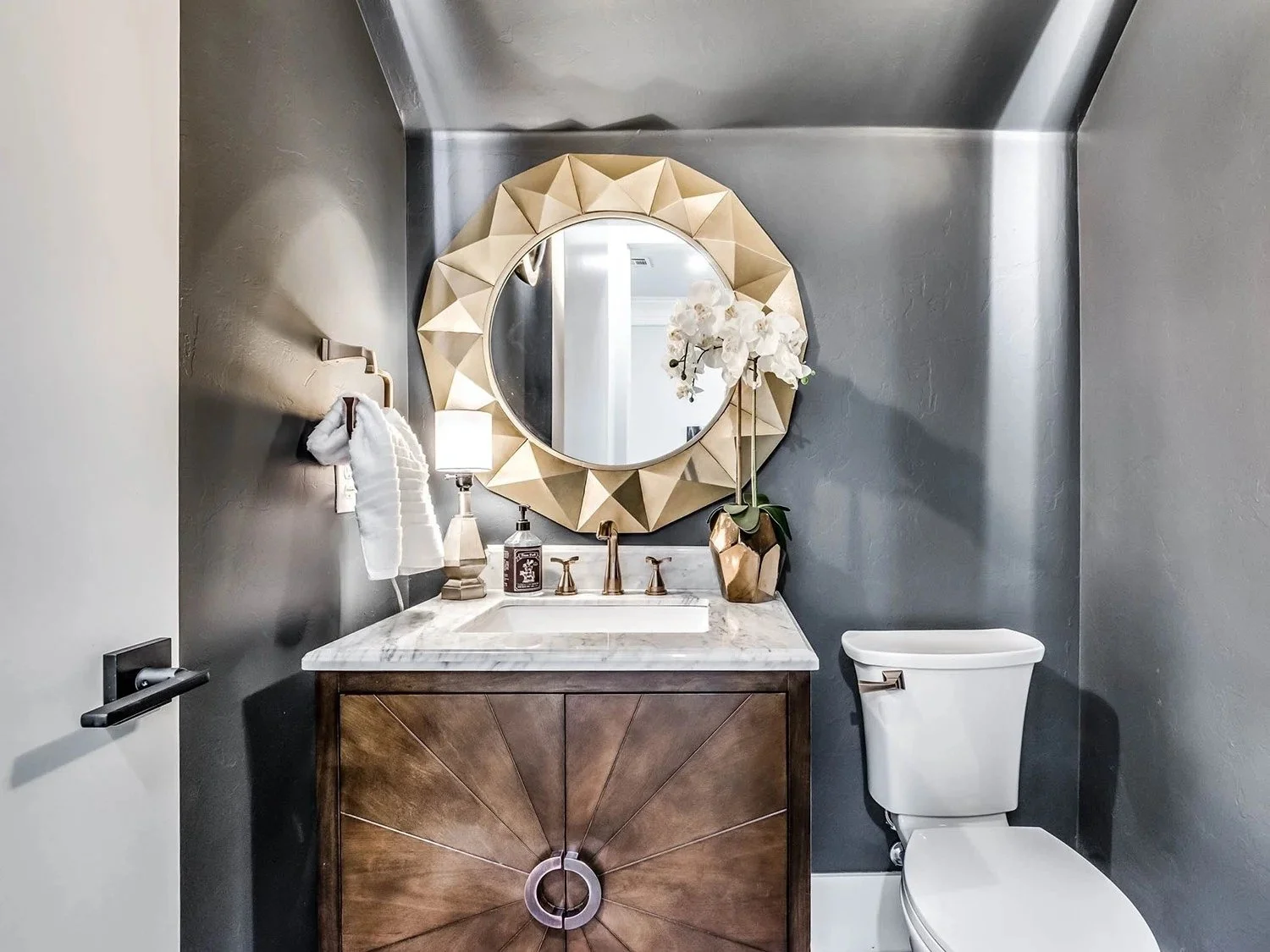 Modern bathroom with a dark gray wall, a round decorative mirror, a marble countertop vanity with a brass faucet, a white orchid in a gold vase, a small table lamp, and a white toilet.