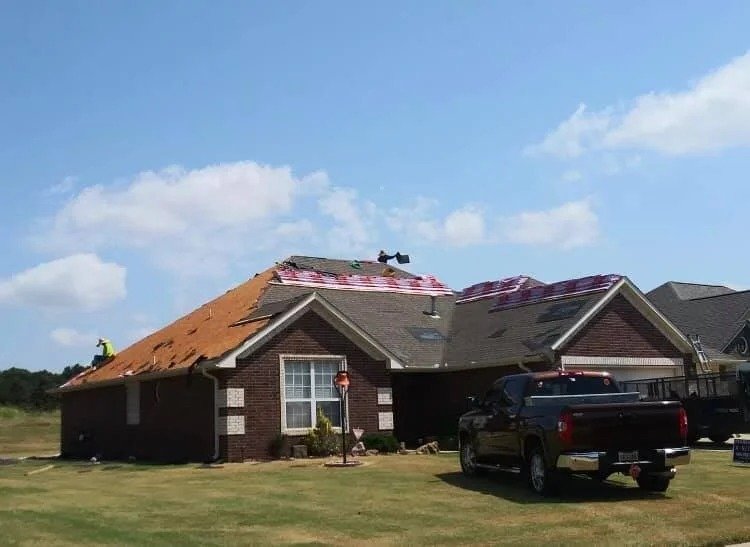 A house under construction with roofing work being done, construction worker on the roof, parked black pickup truck in front, and a partly cloudy sky.