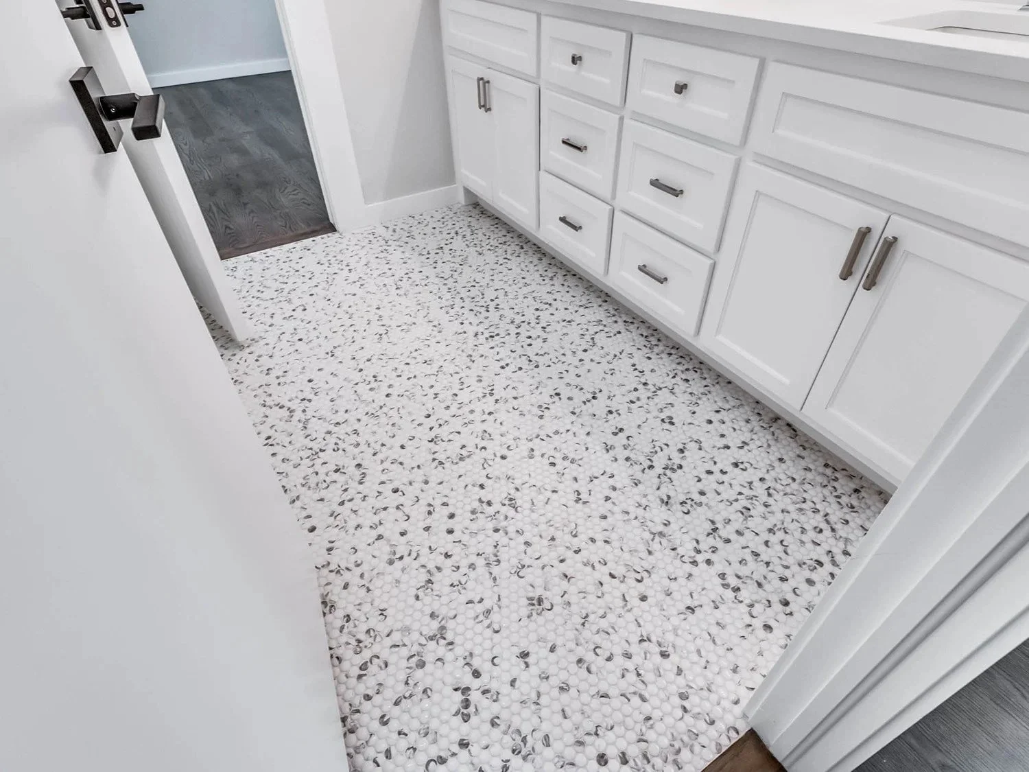 A white bathroom with a terrazzo tile floor, white cabinets with black handles, and a partially open white door leading to a room with dark hardwood floors.