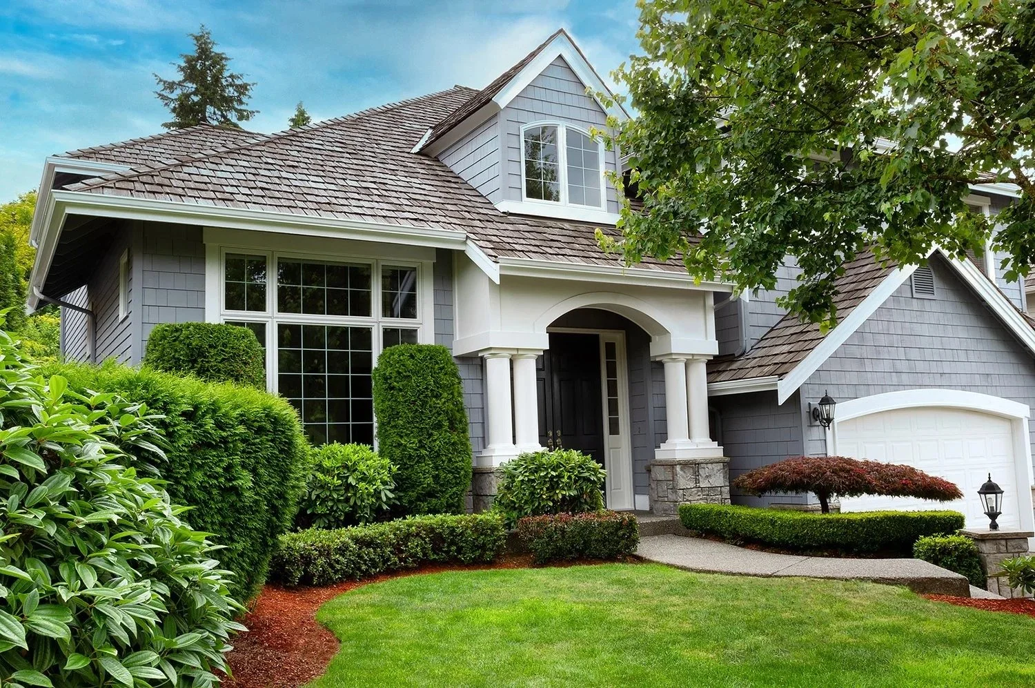 A two-story house with gray siding, white trim, and a brown shingle roof, surrounded by lush green landscaping including bushes, trees, and a well-maintained lawn.
