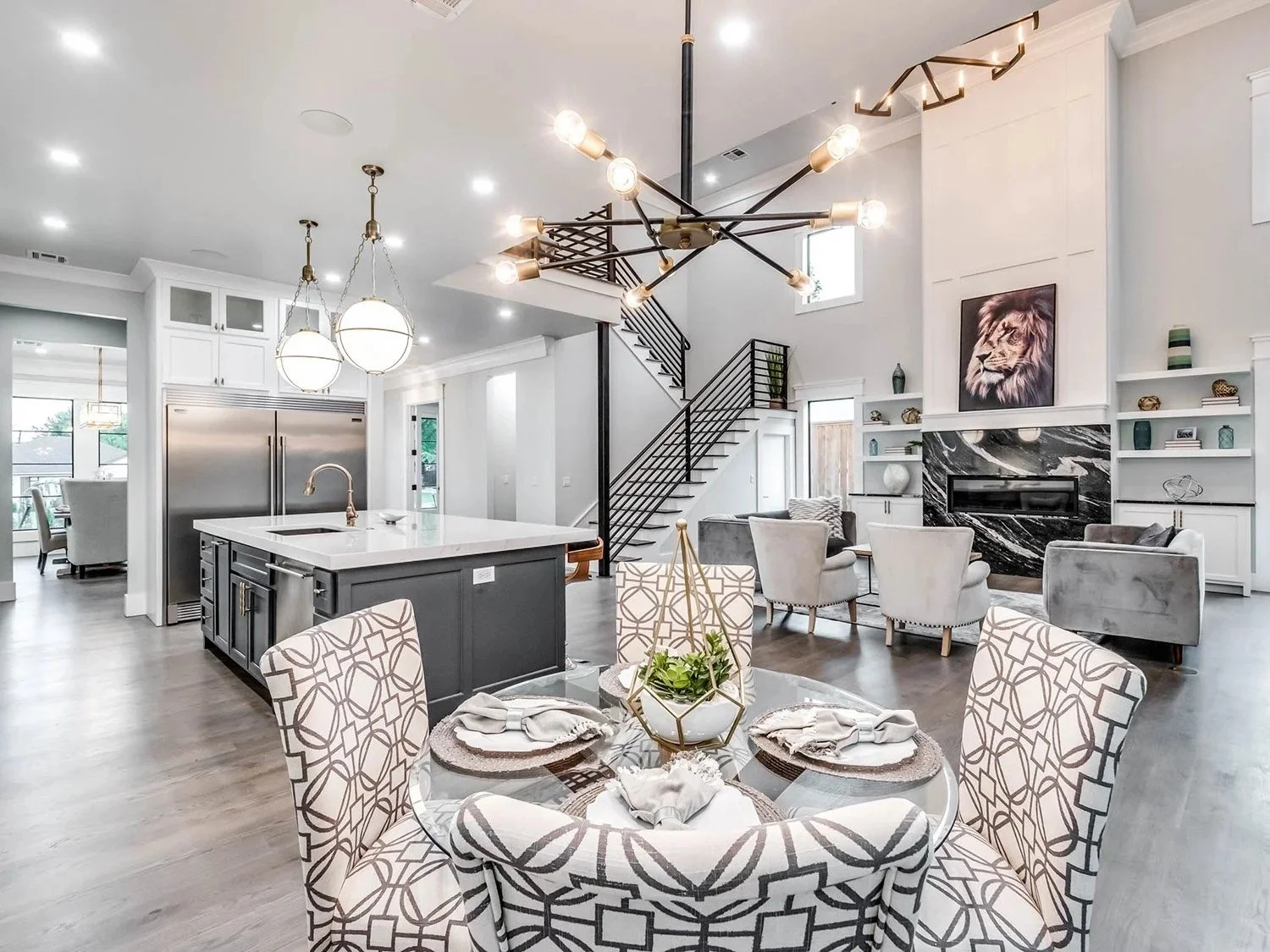 Open-concept living room and kitchen with white walls, hardwood floors, modern furniture, a black and gold chandelier, a fireplace with black marble surround, and a staircase with black railing.