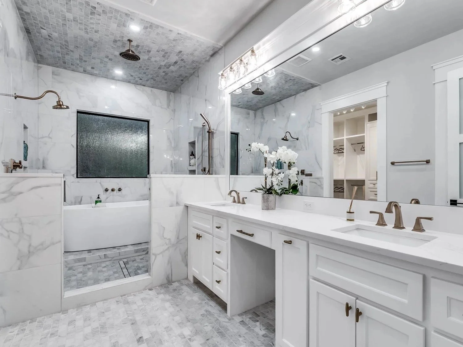 Bright modern bathroom with marble walls, double vanity with brass fixtures, large mirror, and white cabinets. Features walk-in shower with glass partition and bathtub with a frosted window.