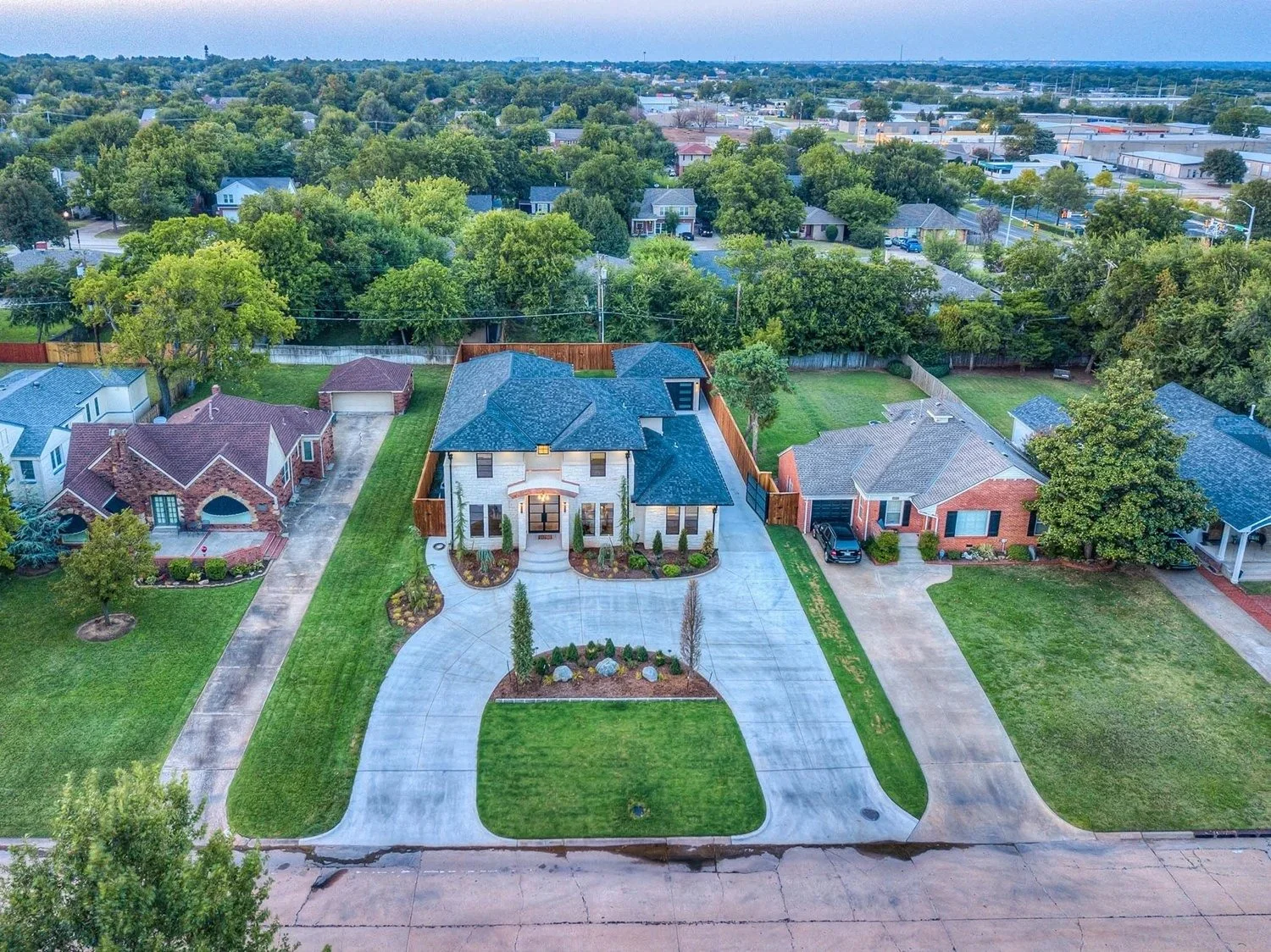 Aerial view of a modern two-story house with a spacious driveway, landscaped front yard, and neighboring houses in a suburban neighborhood surrounded by trees.