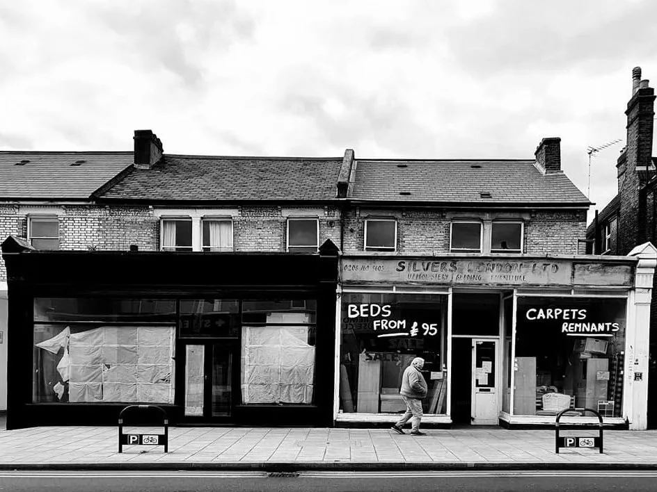 Black and white photo of a strip mall with furniture store and residential buildings above, one person walking on the sidewalk.