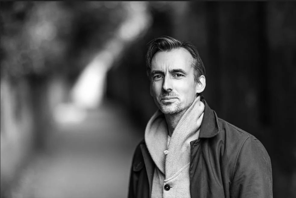 A black and white portrait of a middle-aged man with styled hair, wearing a hoodie under a jacket, standing outdoors with a blurred background.