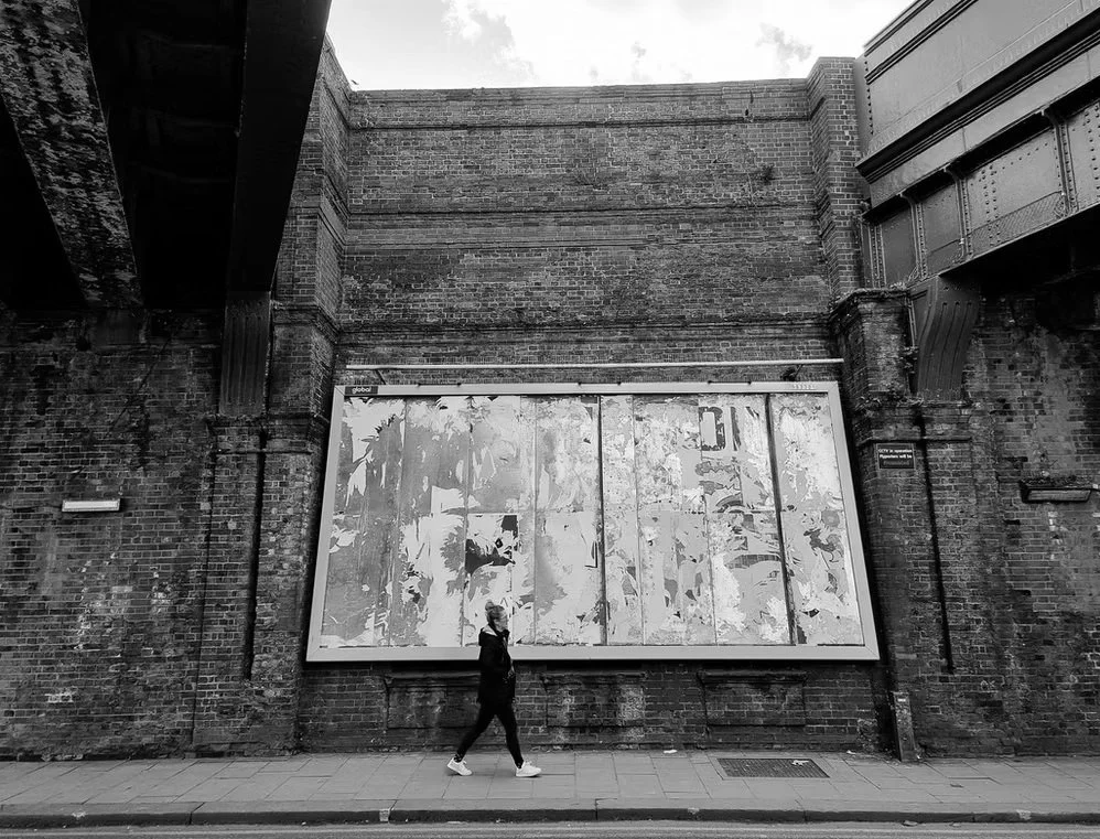 A person walking past a large, weathered billboard mounted on a brick wall under a bridge in an urban setting.