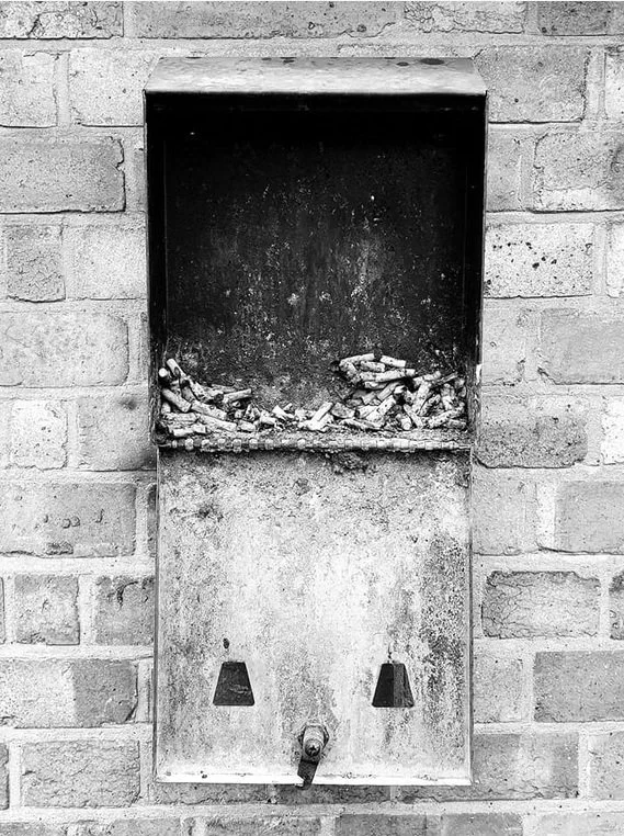 An outdoor metal trash receptacle attached to a brick wall, with a bottom compartment containing a cigarette butt and ash, and a top compartment filled with cigarette butts and ash.