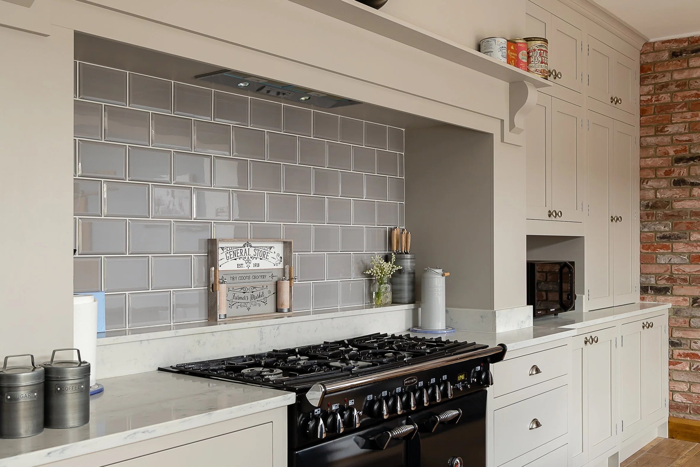 Kitchen with white cabinets, a black stove, and a brick wall on the right side. Grey tiled backsplash and countertop with containers labeled sugar and coffee, a paper towel, and decorative items on the counter.