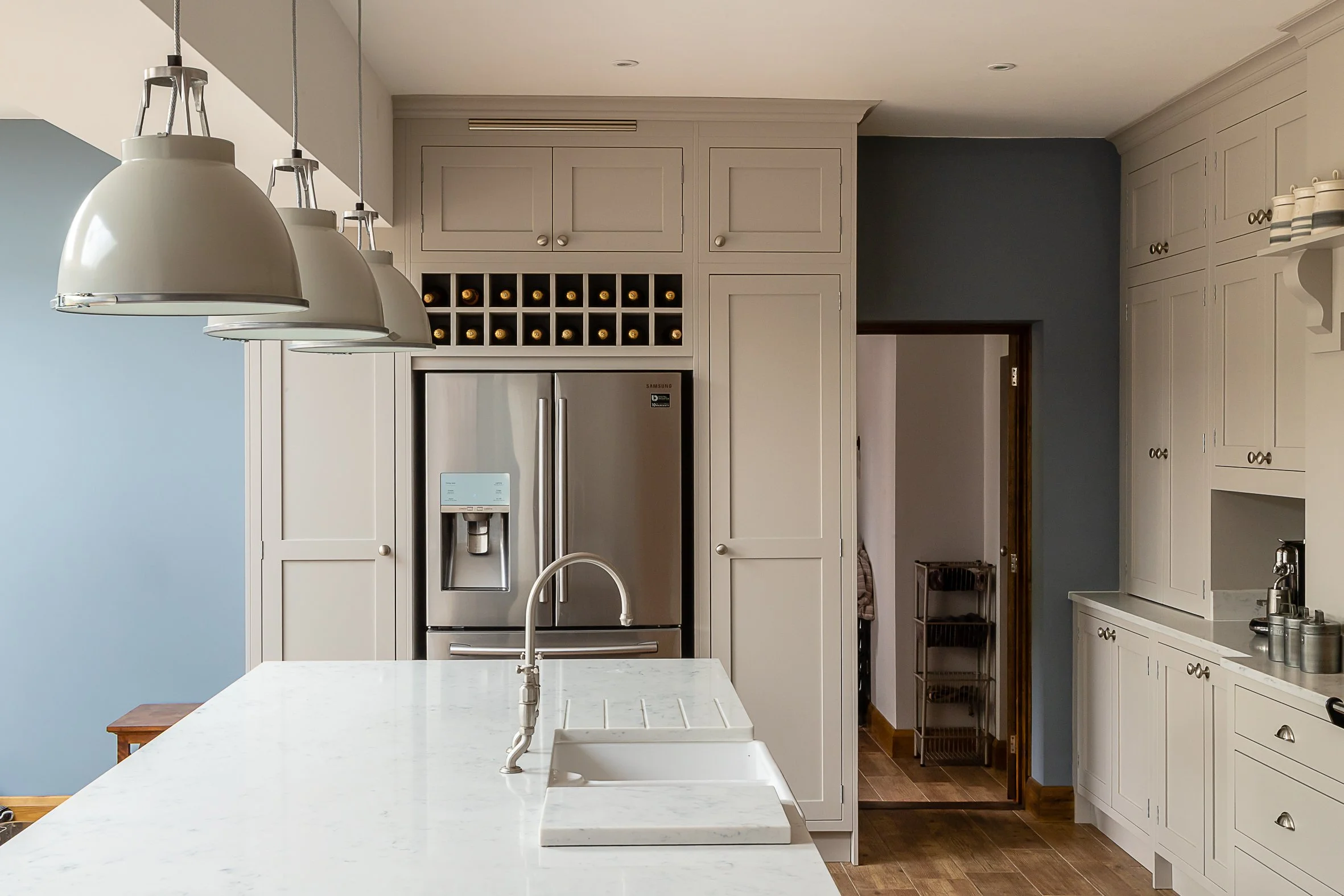 A modern kitchen with white cabinetry, a stainless steel refrigerator, a marble island with a sink, and three white pendant lights hanging above.