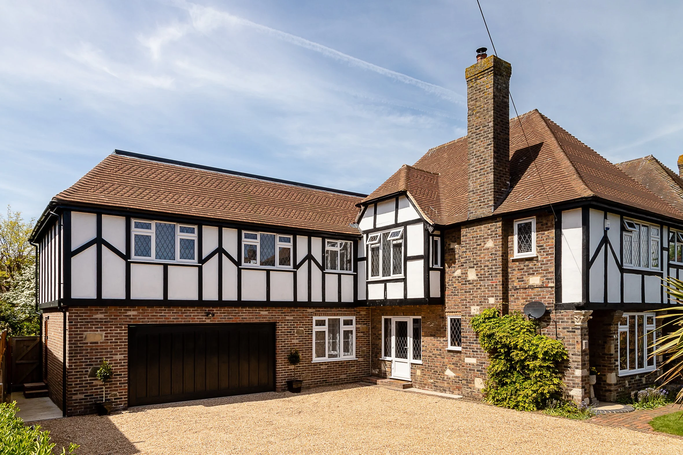 A large two-story house with a brick and timber-framed exterior, brown tiled roof, multiple windows, and a black garage door, under a blue sky with clouds and contrails.