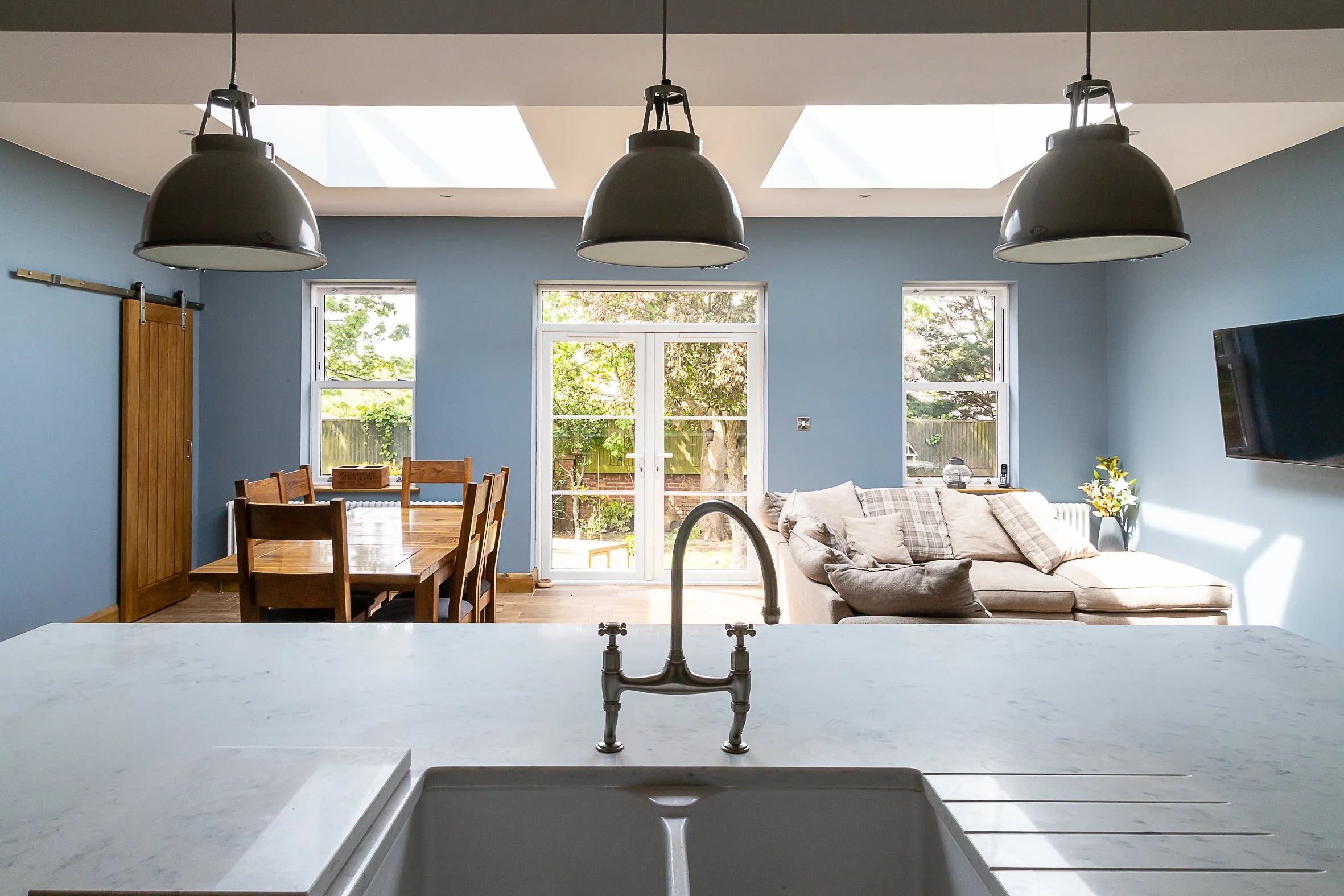 Bright living room with blue walls, large windows, a white sliding door, a beige sofa with cushions, a flat-screen TV, a wooden dining table with chairs, and pendant lights hanging from the ceiling.