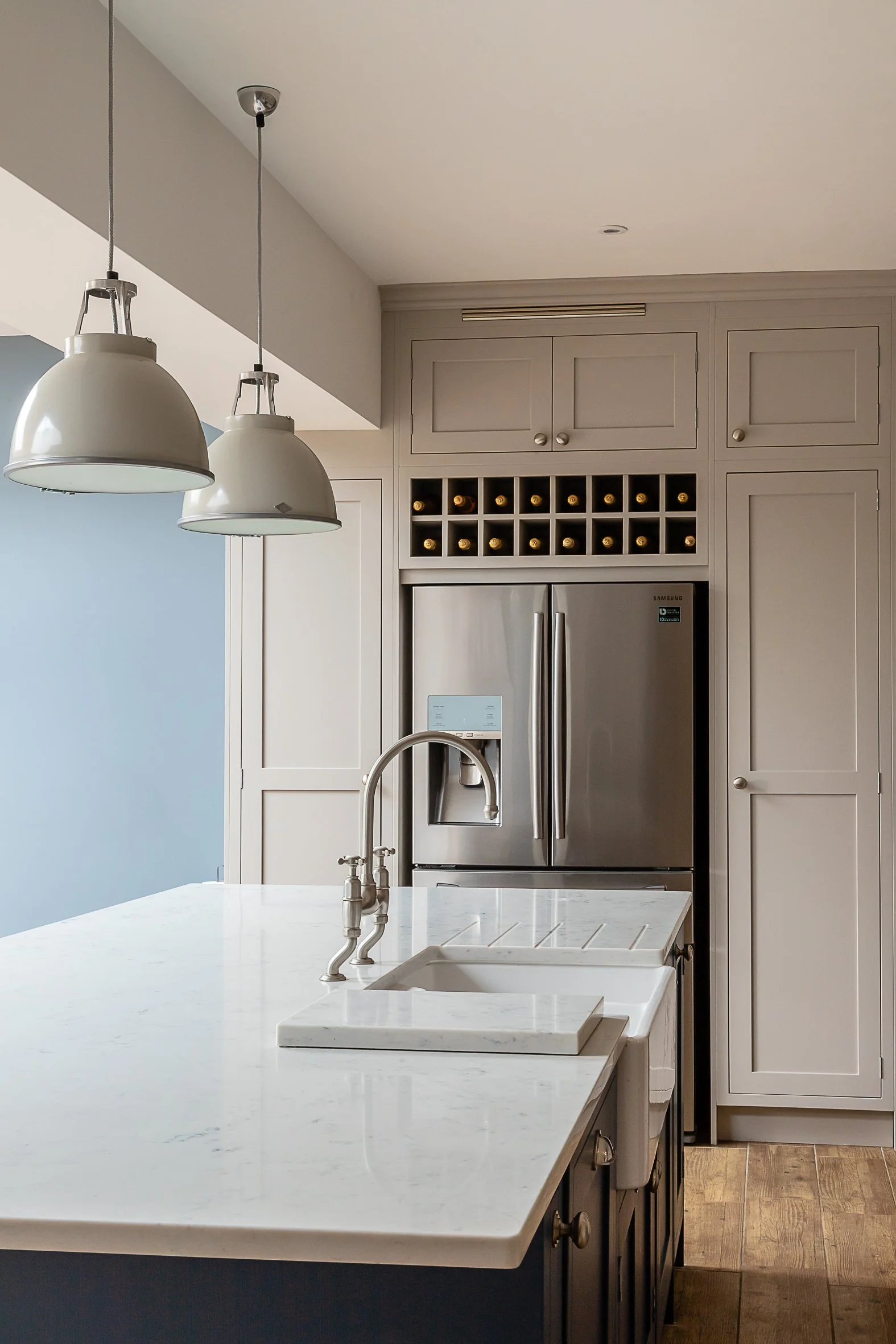 Kitchen with beige cabinets, stainless steel refrigerator, and a white marble island with a sink. Hanging pendant lights above the island and a wall with a wine bottle storage compartment.