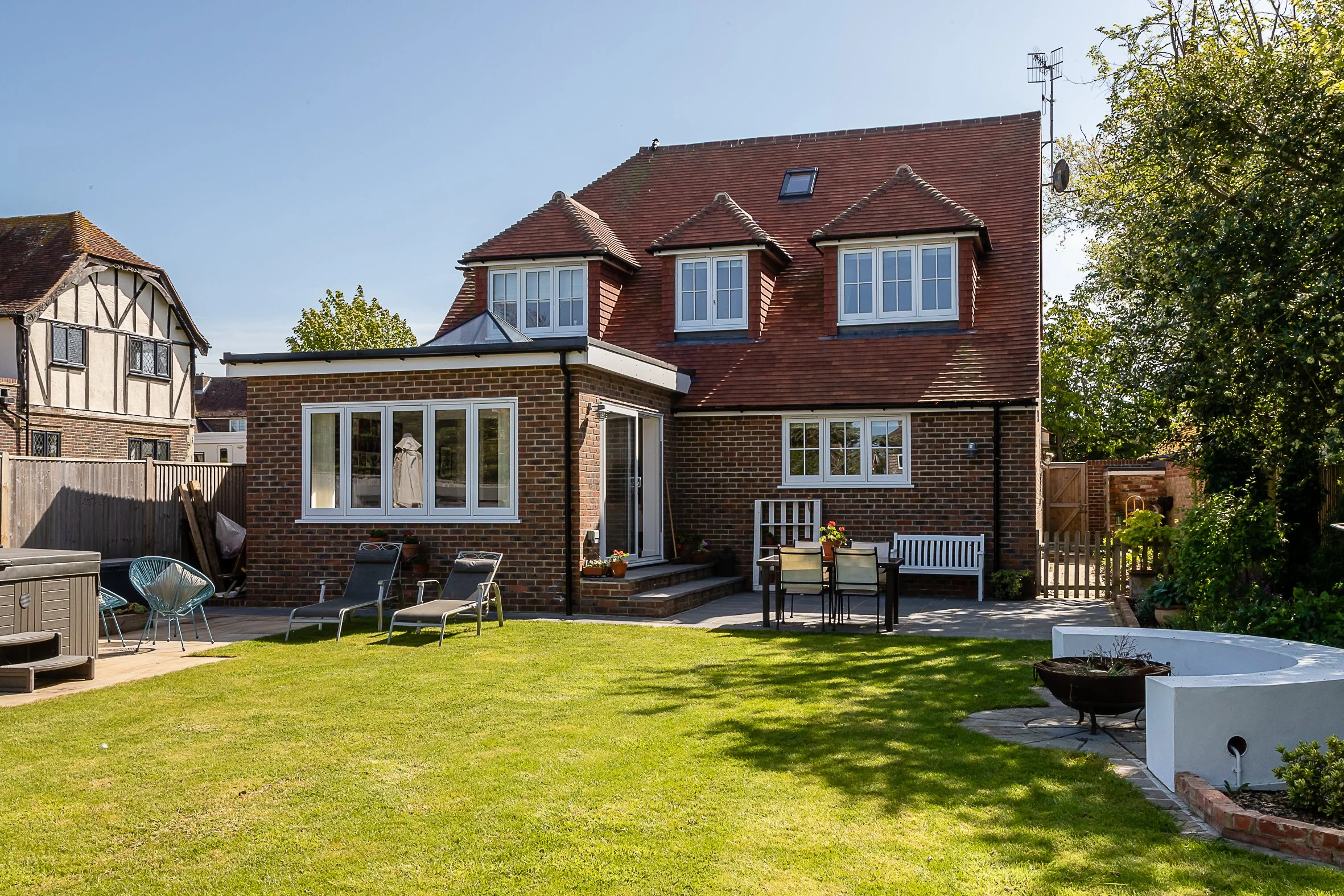 A backyard view of a brick house with multiple windows, a red-tiled roof, and an outdoor patio area with lounge chairs, a table with chairs, and a white bench. The yard is grassy with some trees, garden beds, and a clear blue sky.