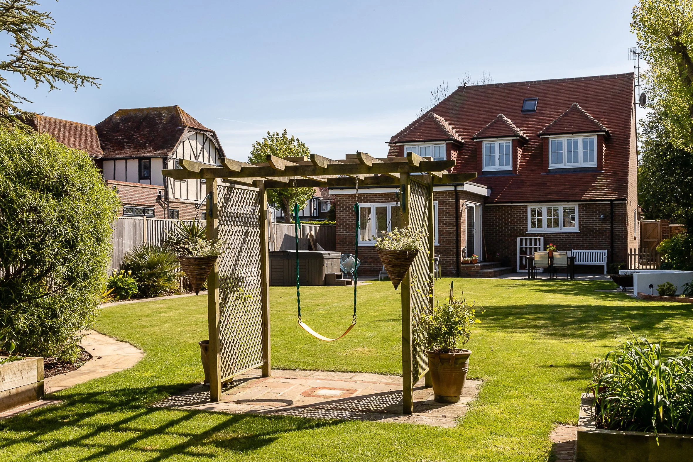 Backyard with a brick house, lawn, outdoor furniture, and a wooden pergola with hanging planters and a swing.